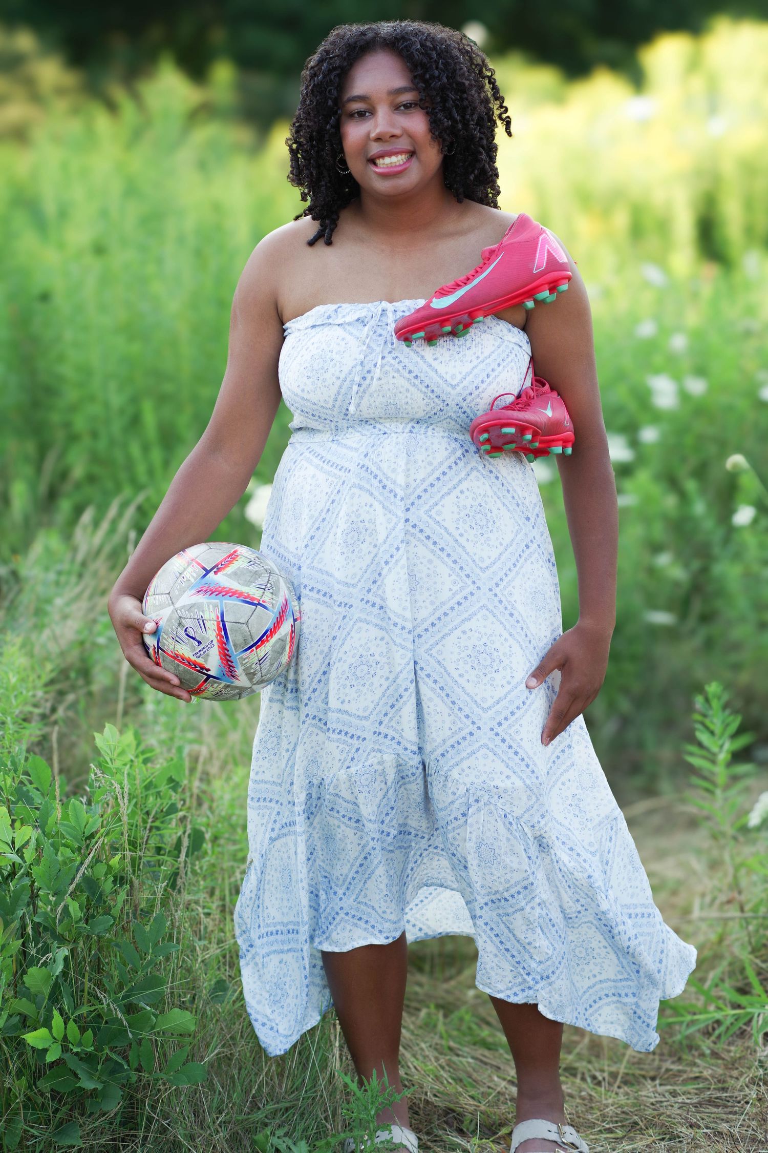 Person in a white patterned summer dress stands in a grassy field holding volleyball and red cleats during outdoor photoshoot.