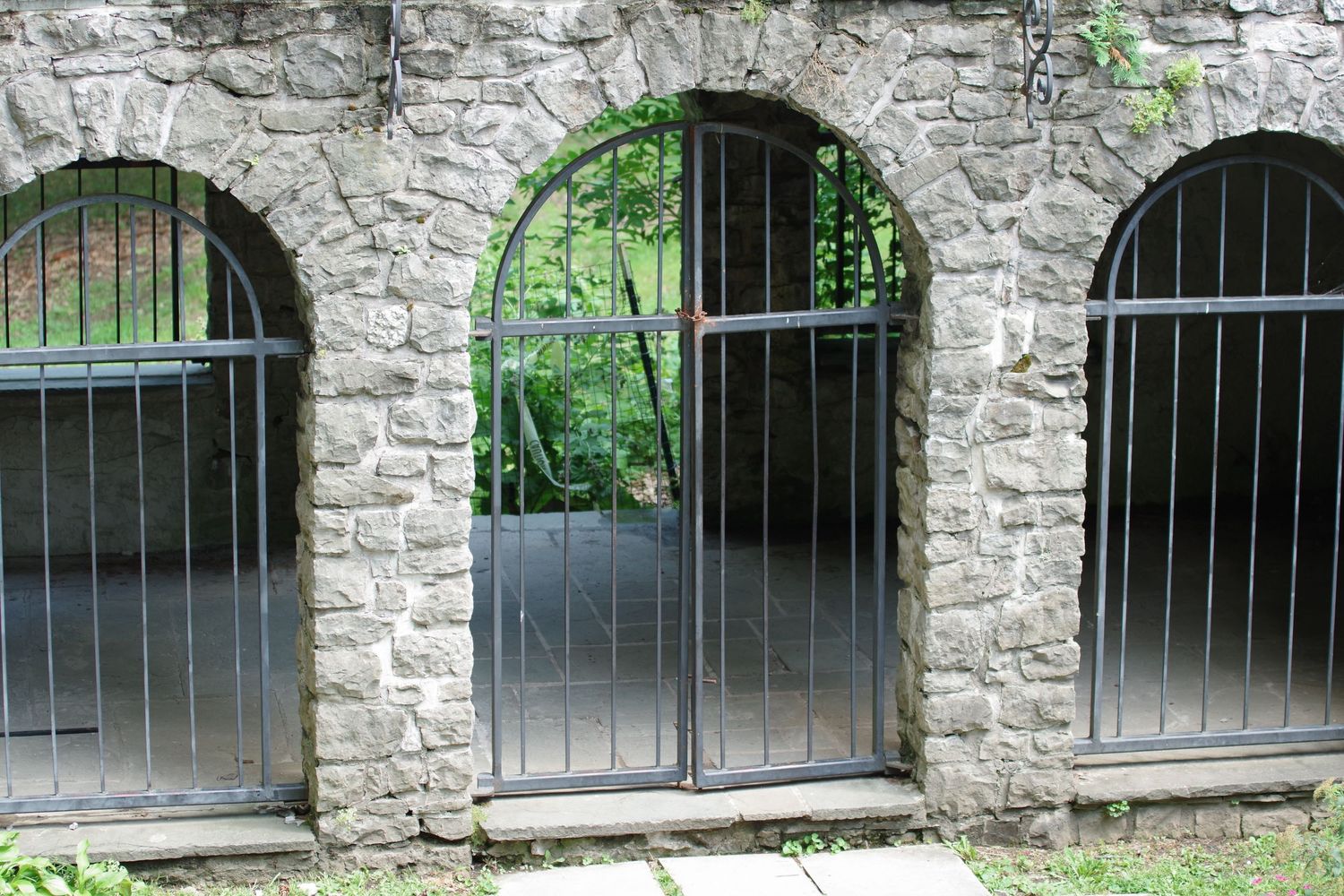 Three arched stone windows with metal bars overlooking green foliage.