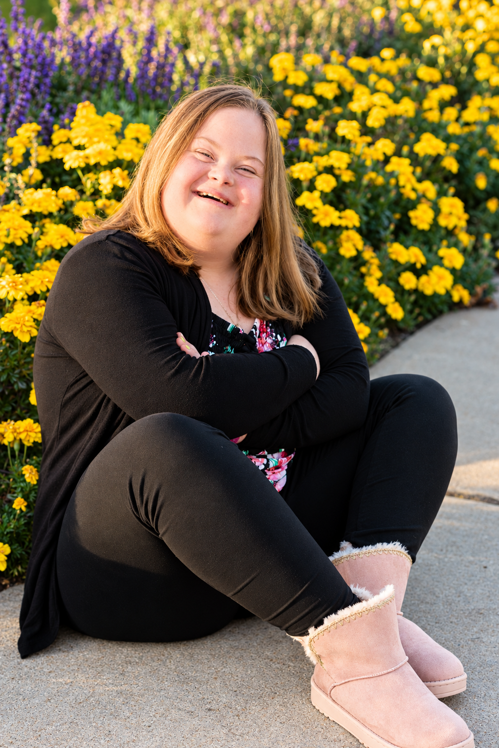 Smiling young woman with Down syndrome sits cross-legged by yellow flowers, wearing black outfit and pink UGG boots.