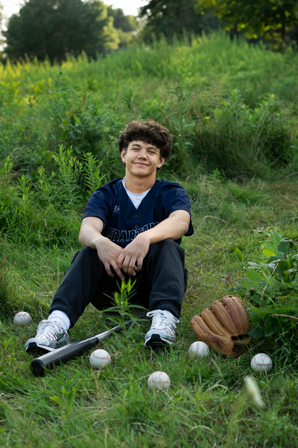 Teen baseball player sitting on grass with bat, glove, and balls, wearing navy jersey, smiling outdoors.