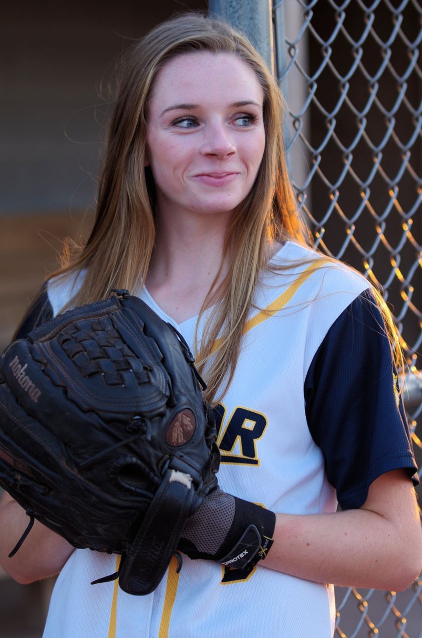 Young female softball player in uniform holding a black fielding glove, smiling near a chain-link fence.