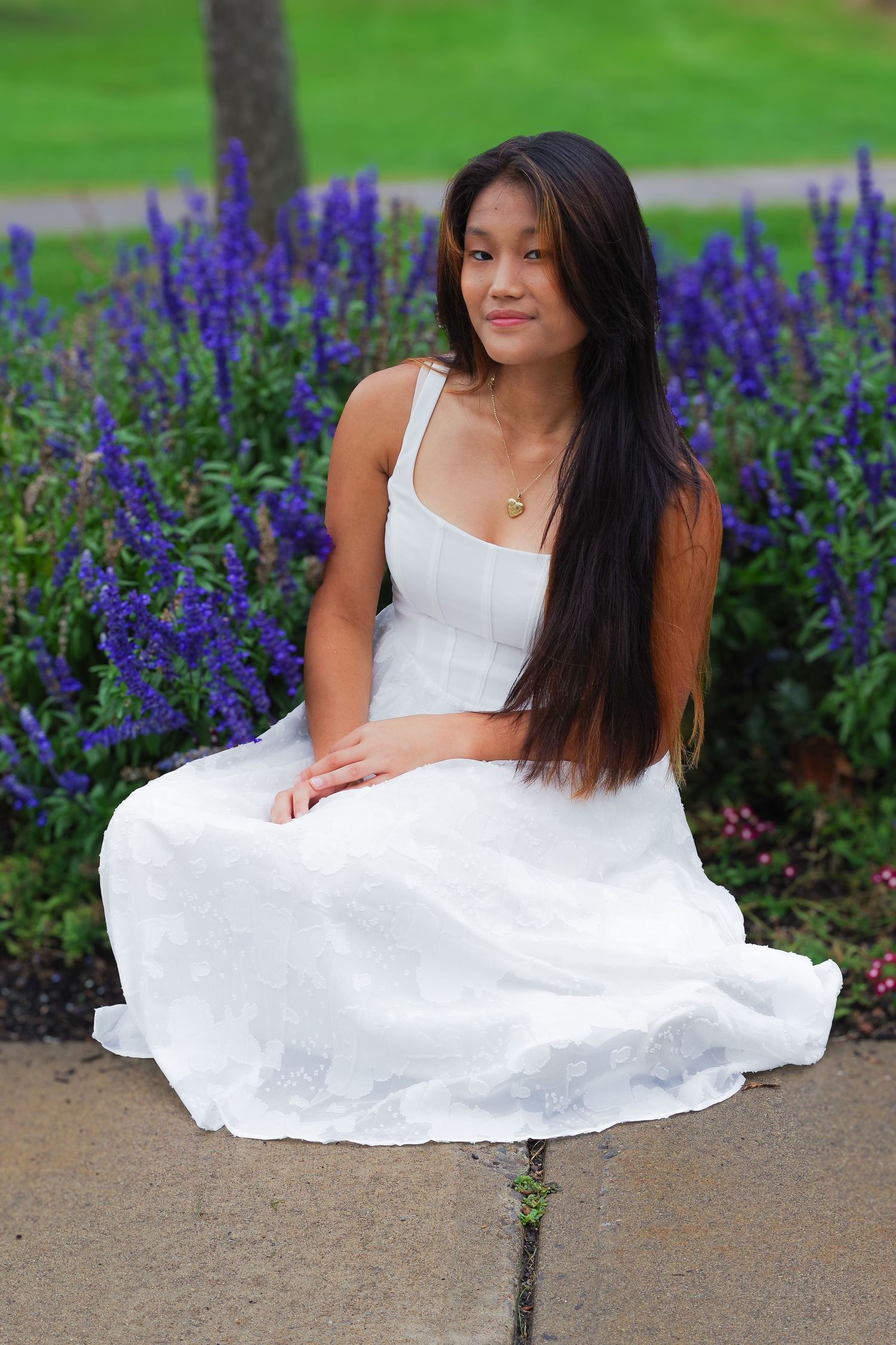 Person in flowing white dress seated among vibrant purple delphiniums in garden.