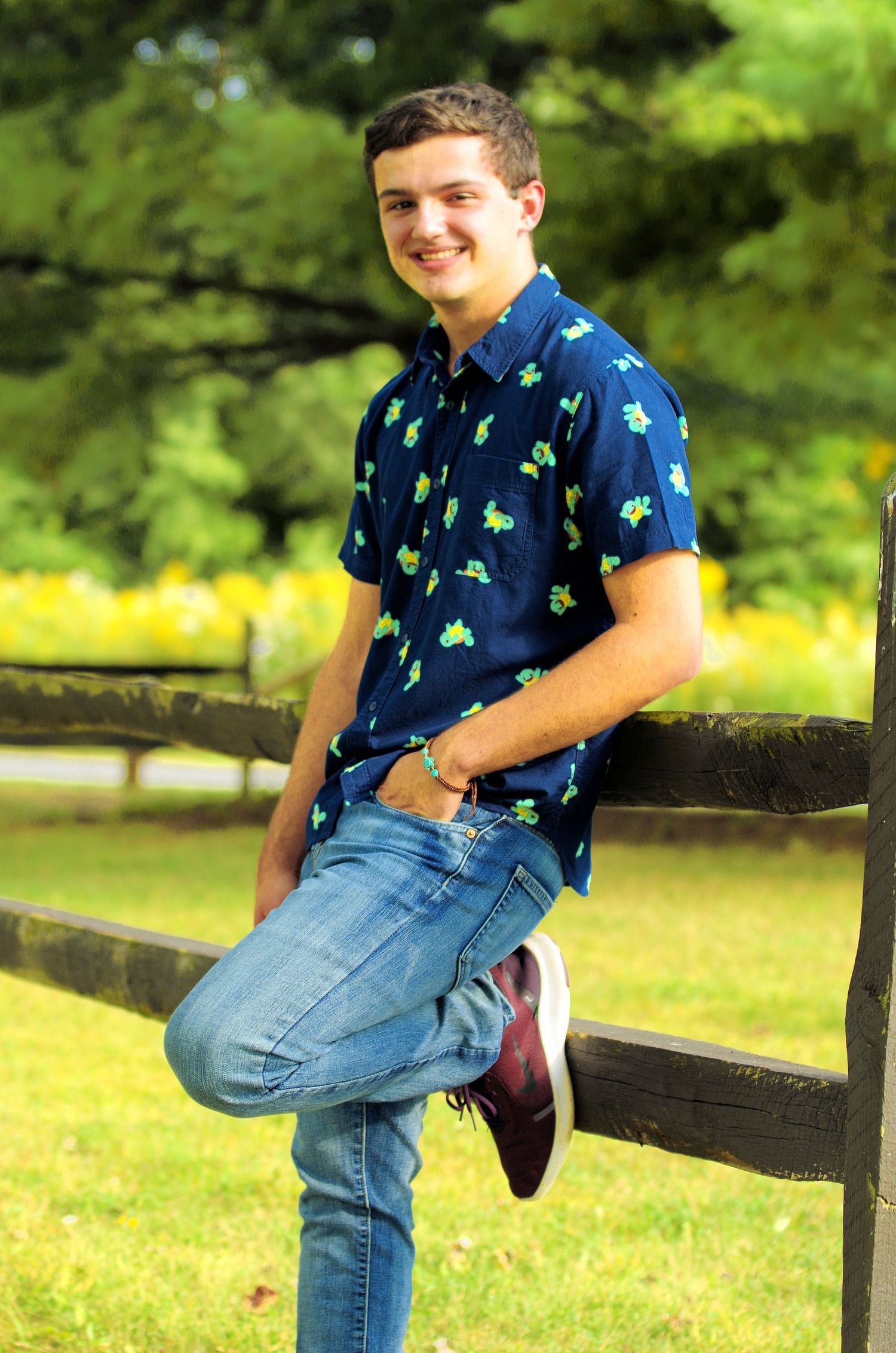 Casual poses on wooden fence in park setting wearing blue patterned shirt and jeans.