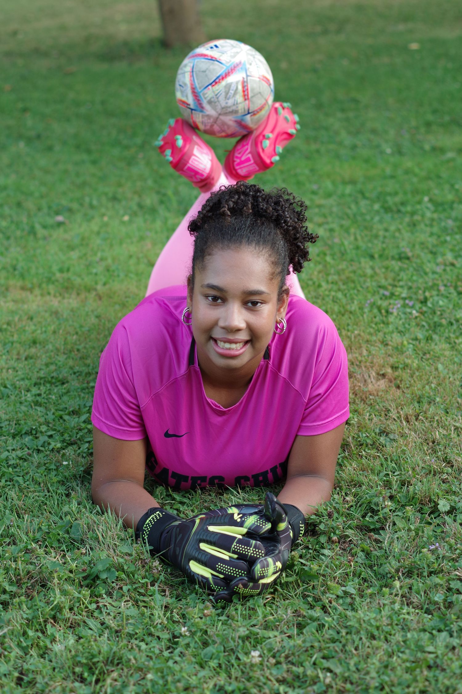 Soccer goalie in pink jersey lying on grass with a ball and gloves.
