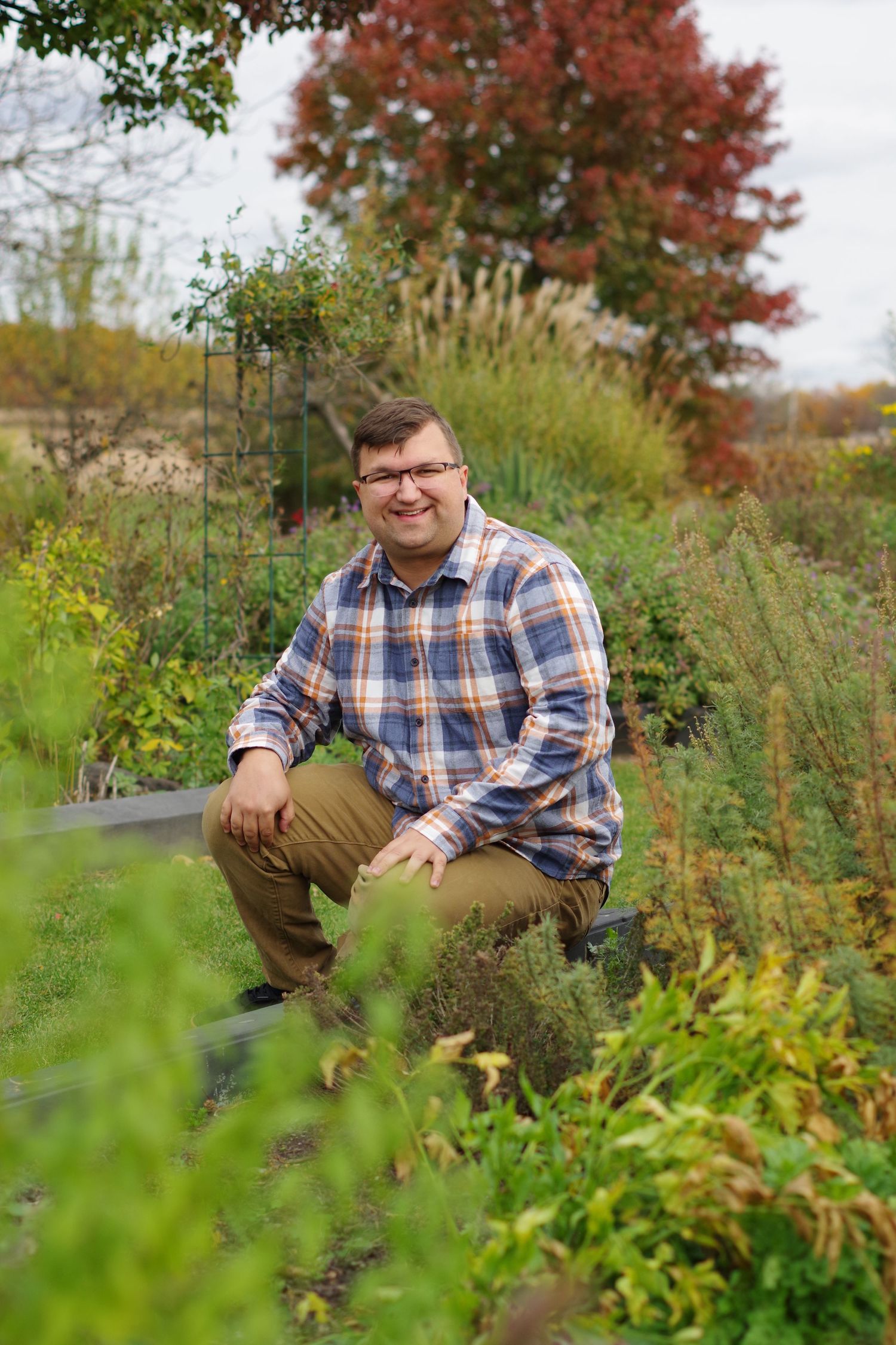 Person in plaid shirt kneeling in lush garden with autumn foliage.