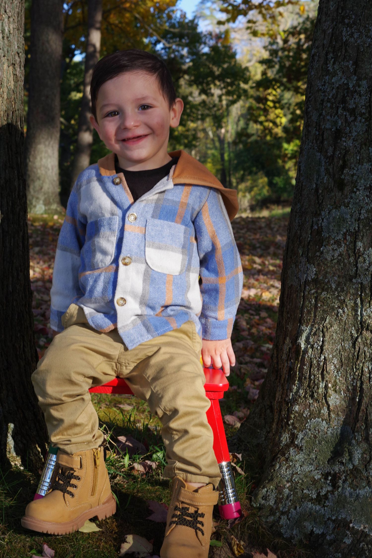 Child wearing striped jacket and khaki pants poses between trees in autumn forest.
