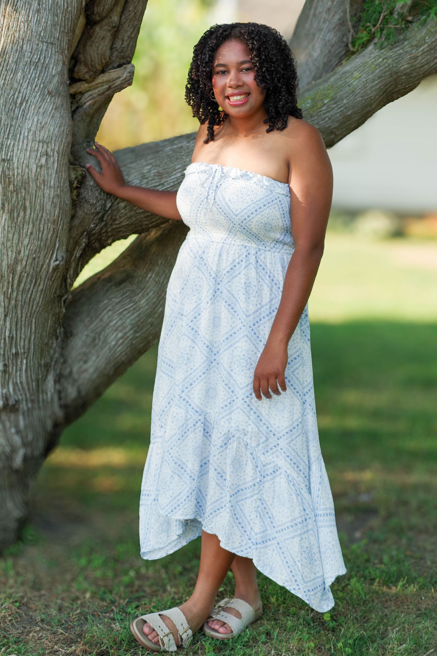A person wearing a flowing white eyelet dress poses gracefully near a tree trunk in a sunlit outdoor setting.