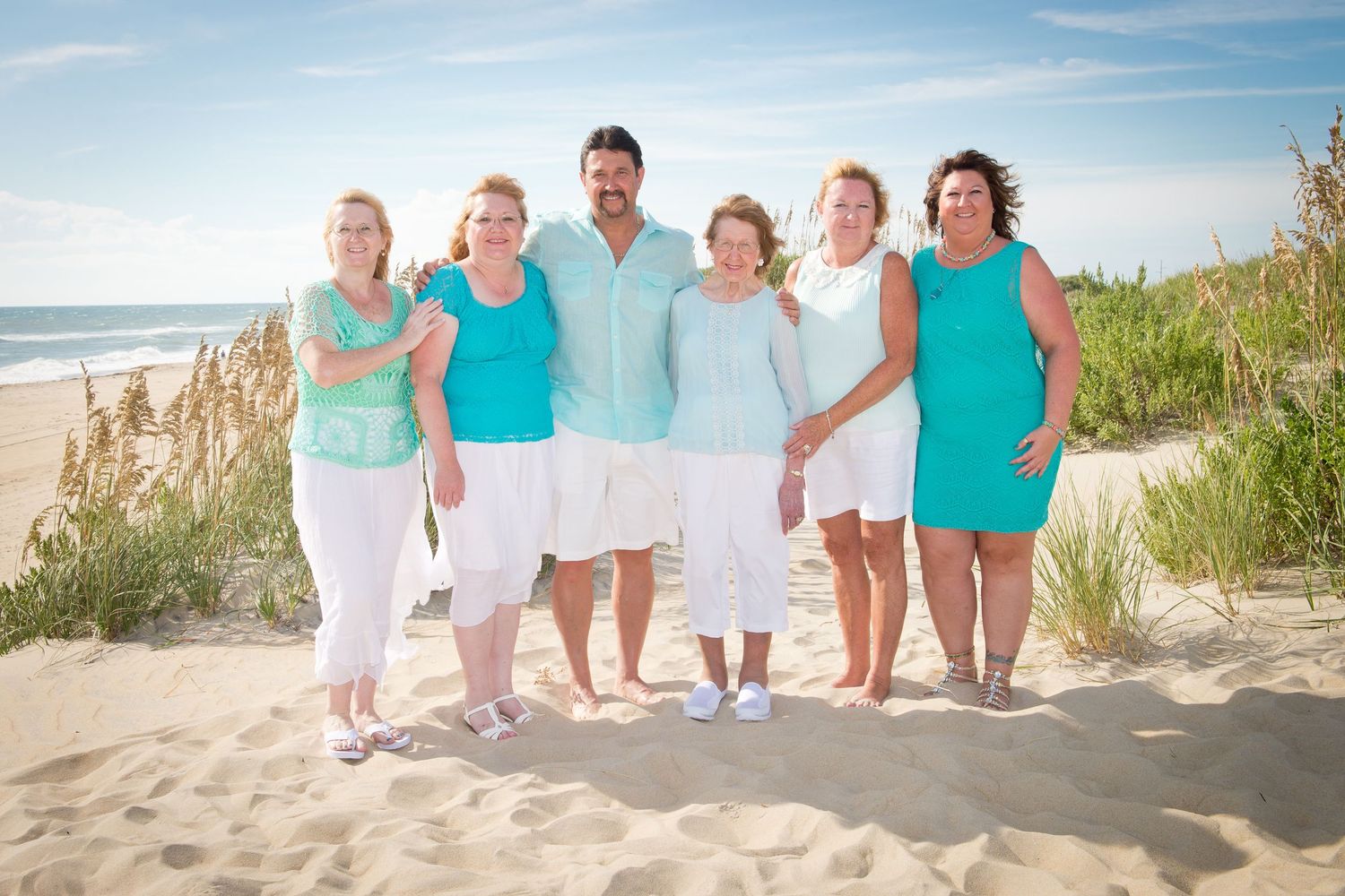 Group wearing coordinating turquoise and white outfits poses together on a sunny beach with sand dunes and ocean view.
