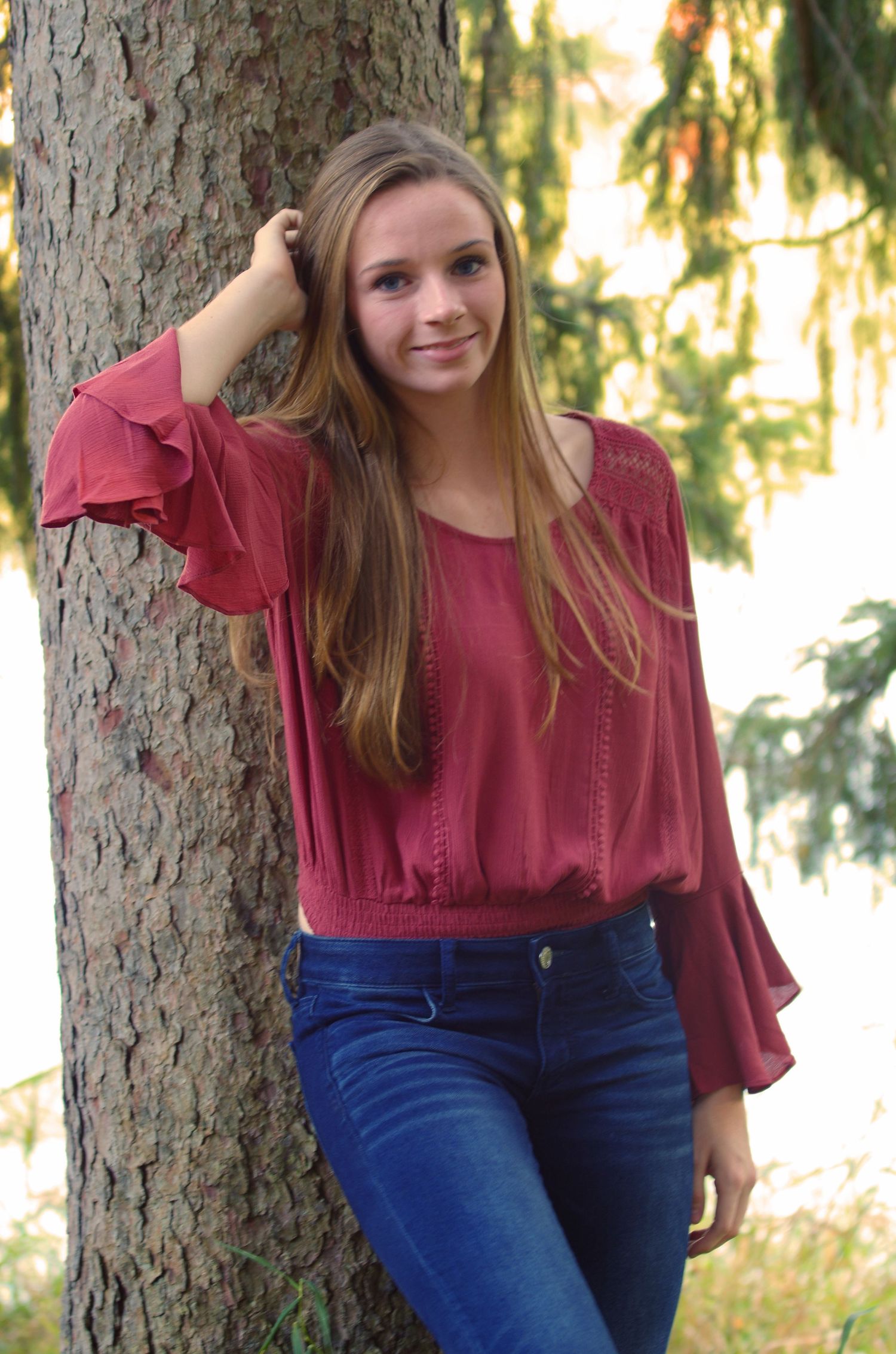 Portrait against tree trunk wearing burgundy top and blue jeans in outdoor setting.