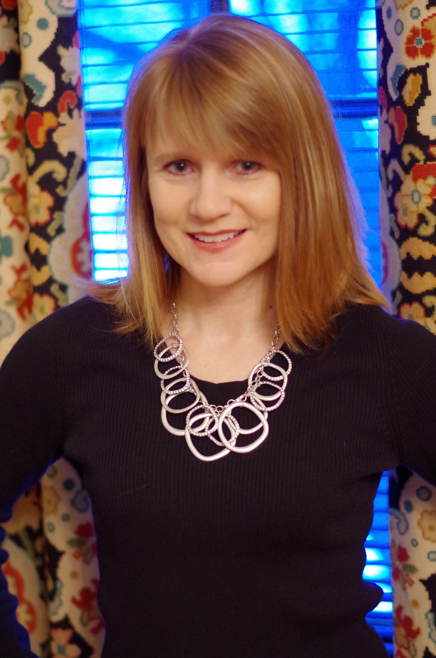 Indoor portrait with black sweater and modern silver circle necklace against blue-lit background.