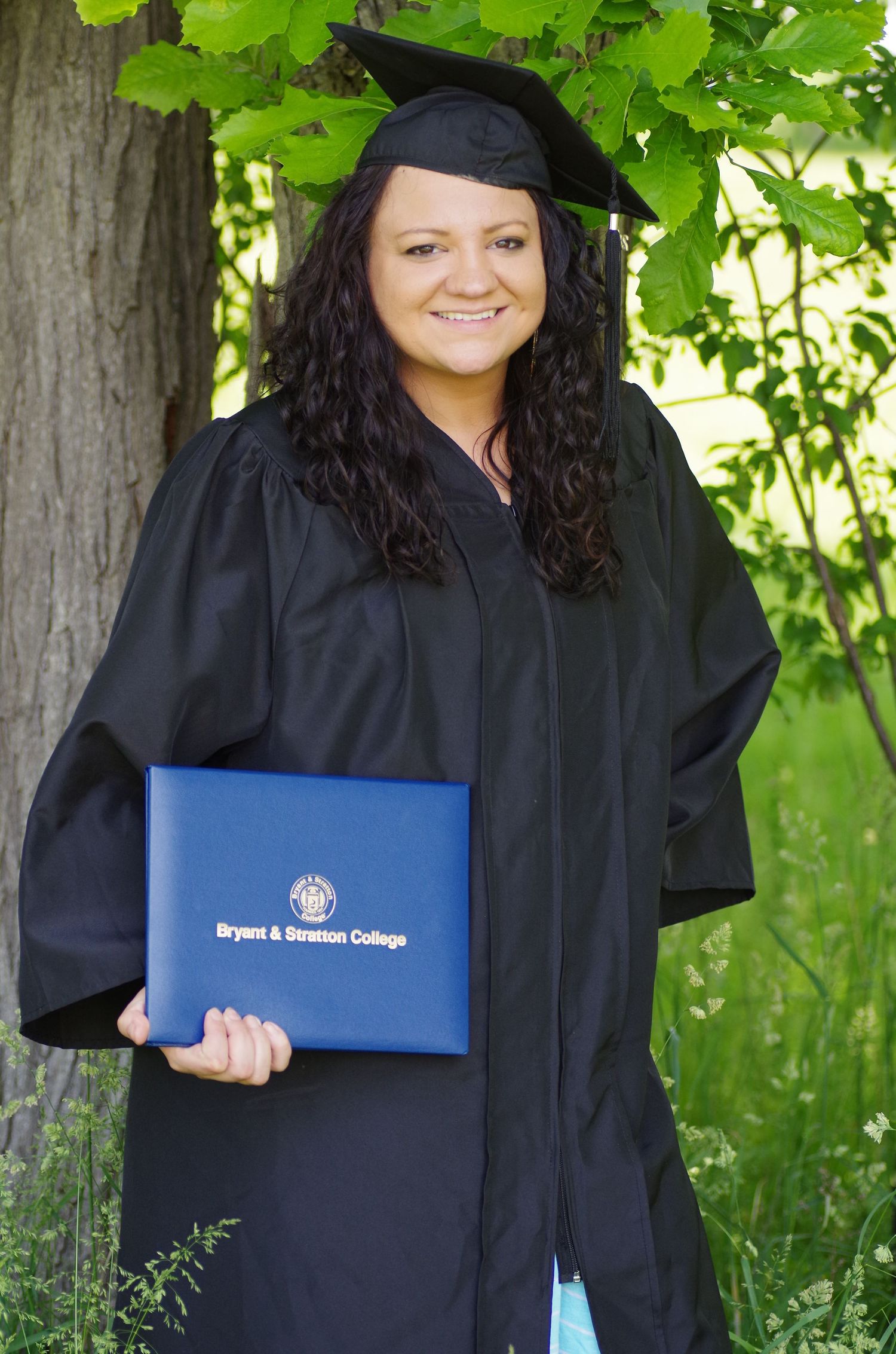 Graduate in black cap and gown holding blue Bryant & Stratton College diploma folder against leafy background.