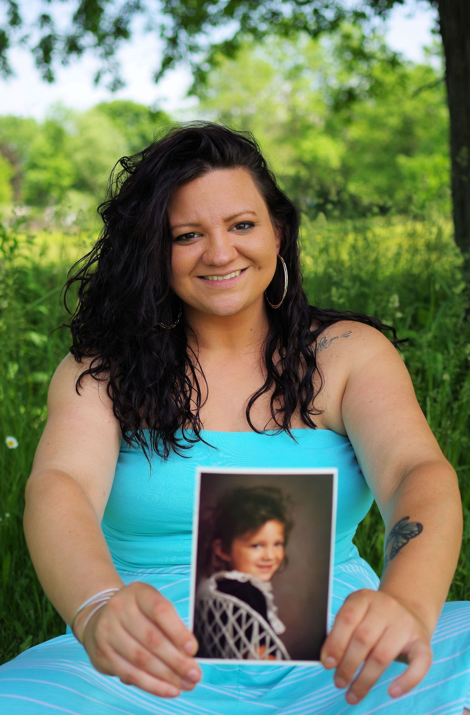 Graduate in turquoise dress holds childhood photos while sitting in a grassy outdoor setting.