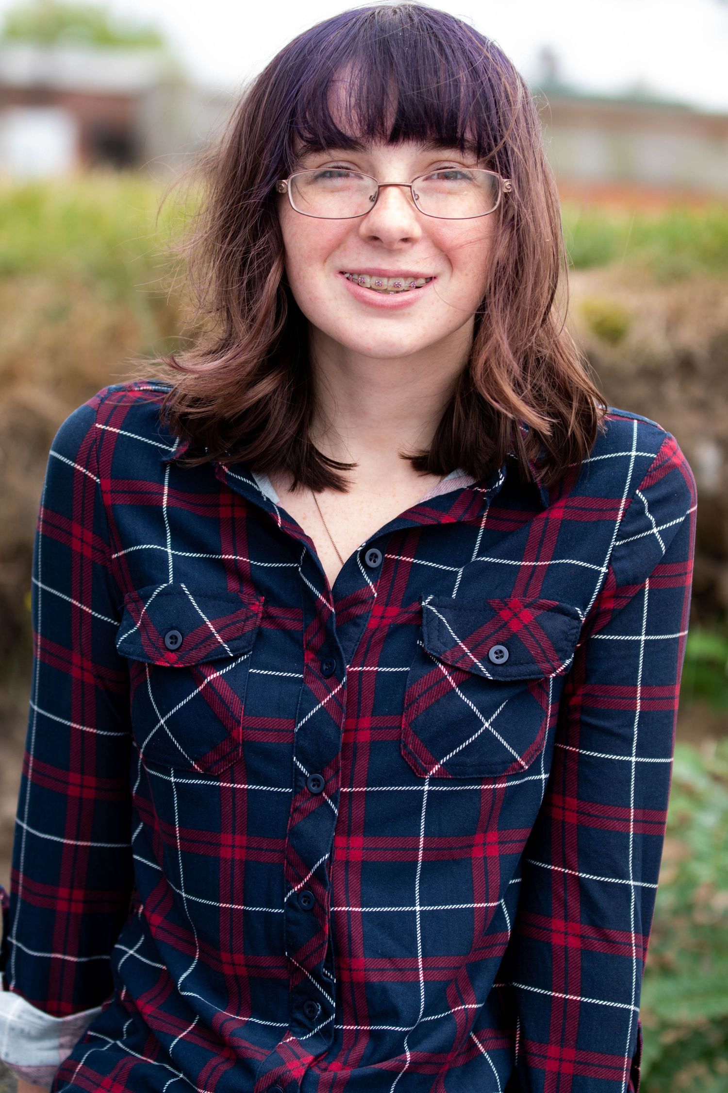 Person wearing glasses and plaid shirt smiles outdoors against blurred background.