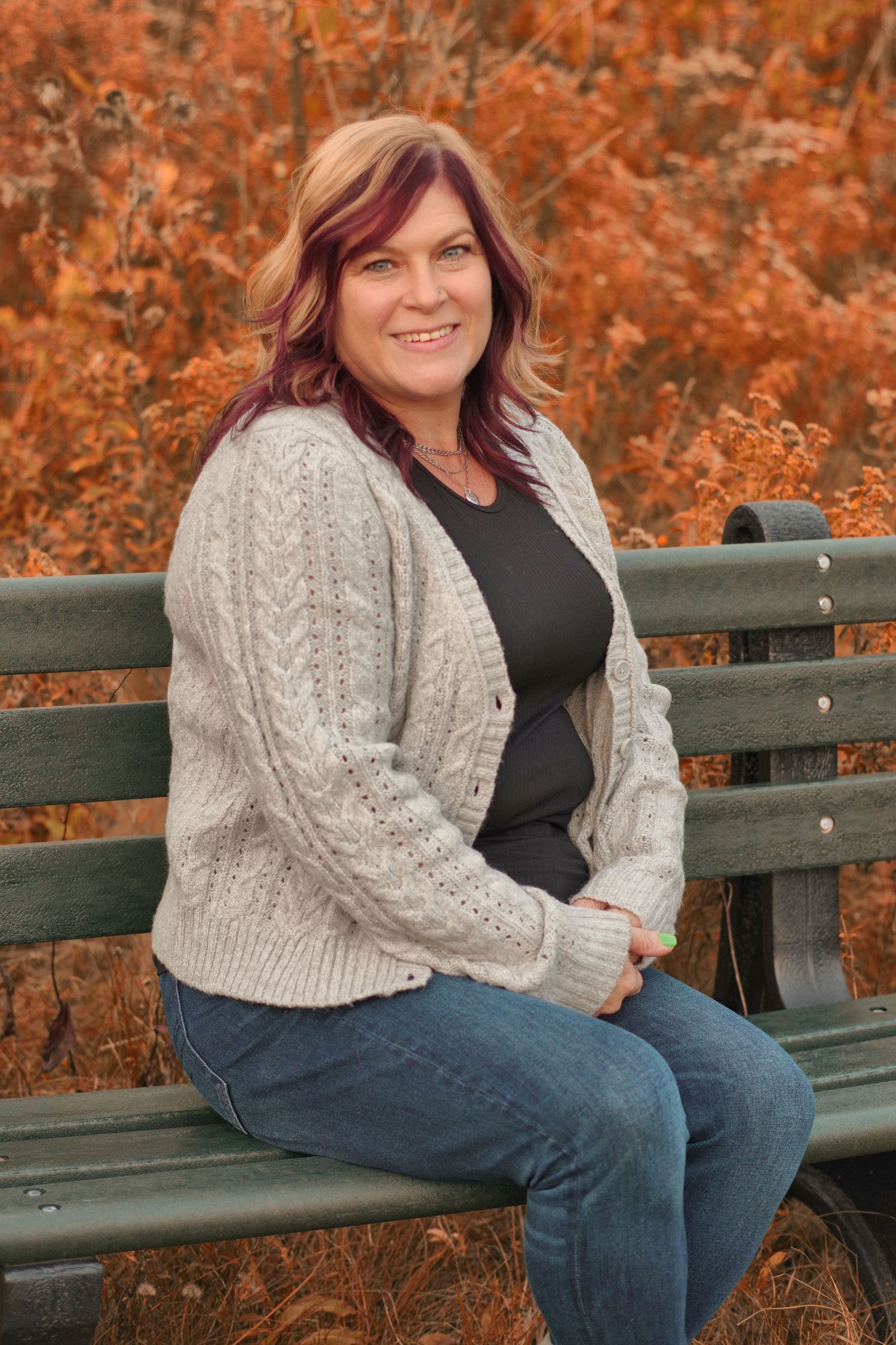 A smiling woman with auburn highlights sits on a green park bench wearing a cream cable-knit cardigan and jeans in autumn.