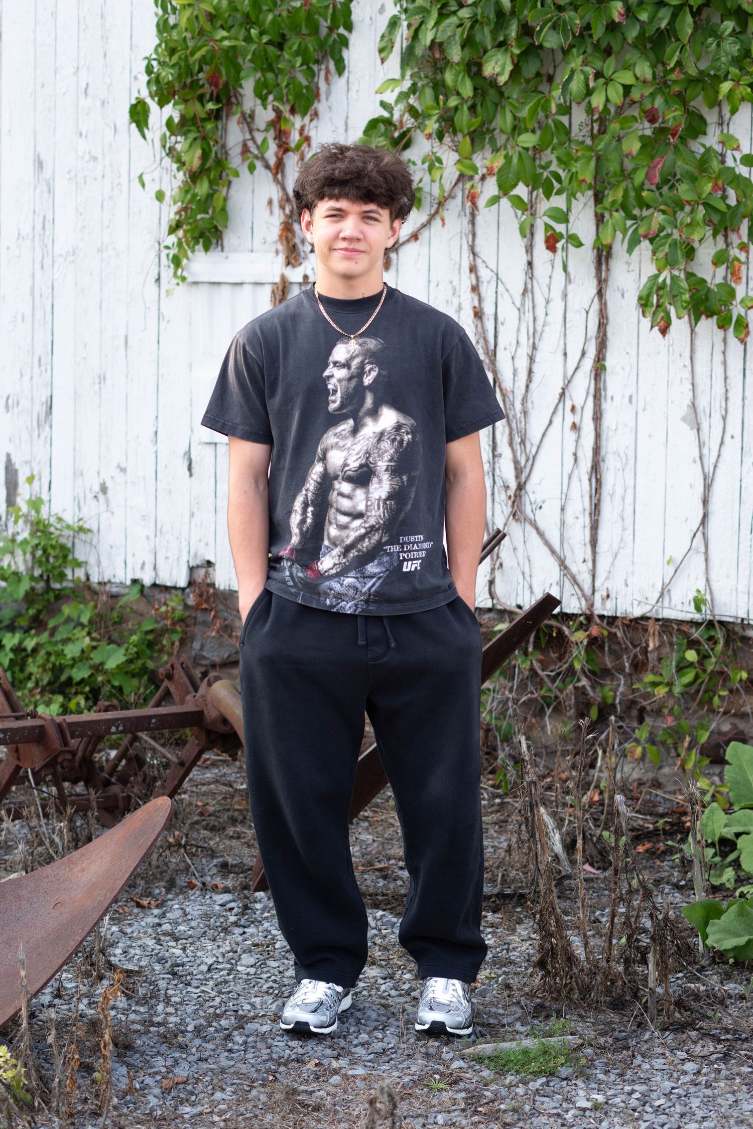 Young man in black graphic tee and sweatpants posing outdoors near rustic white barn wall with ivy.