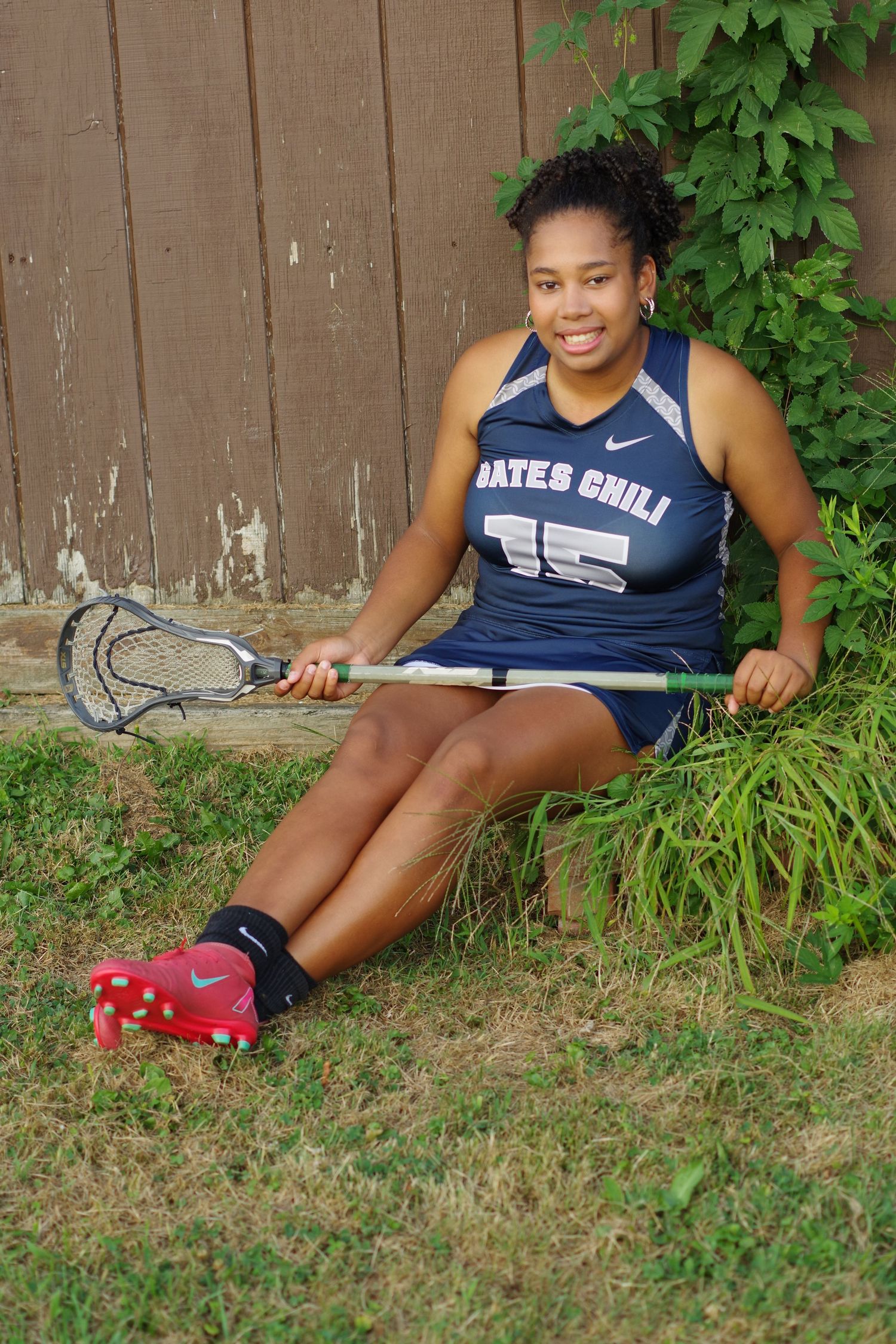 Lacrosse player in navy uniform sits casually with equipment against brown fence.
