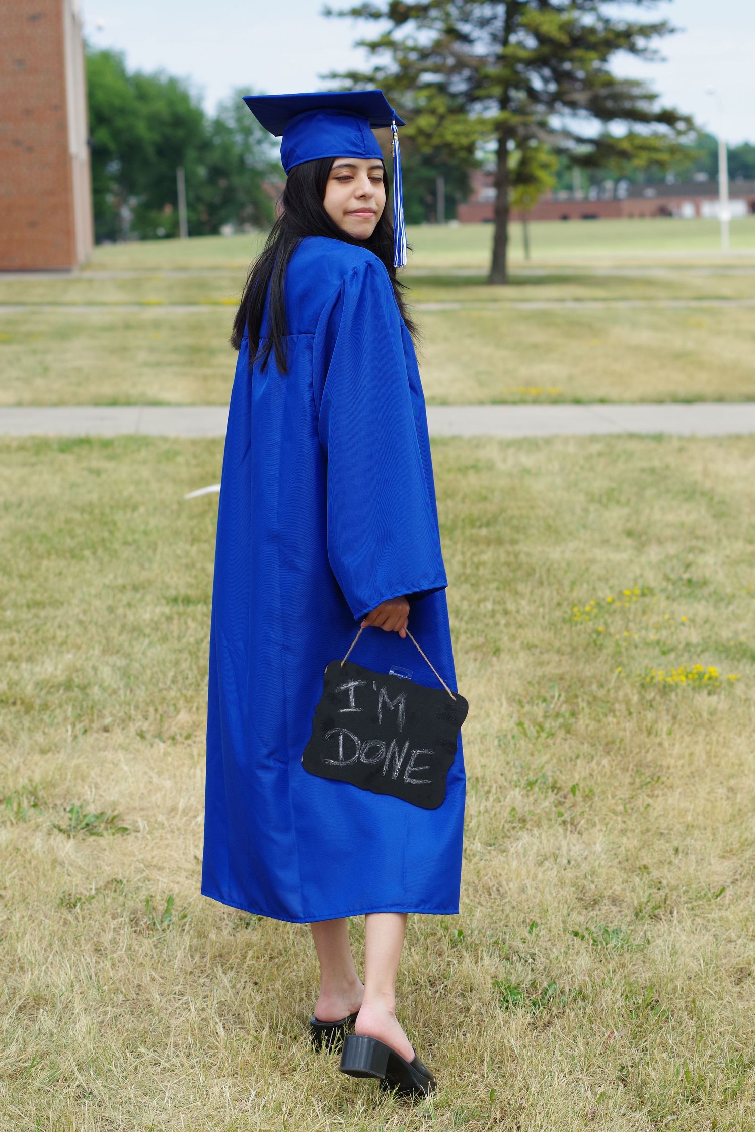 Graduate in blue gown holds 'I'm Done' sign while posing outdoors on campus.