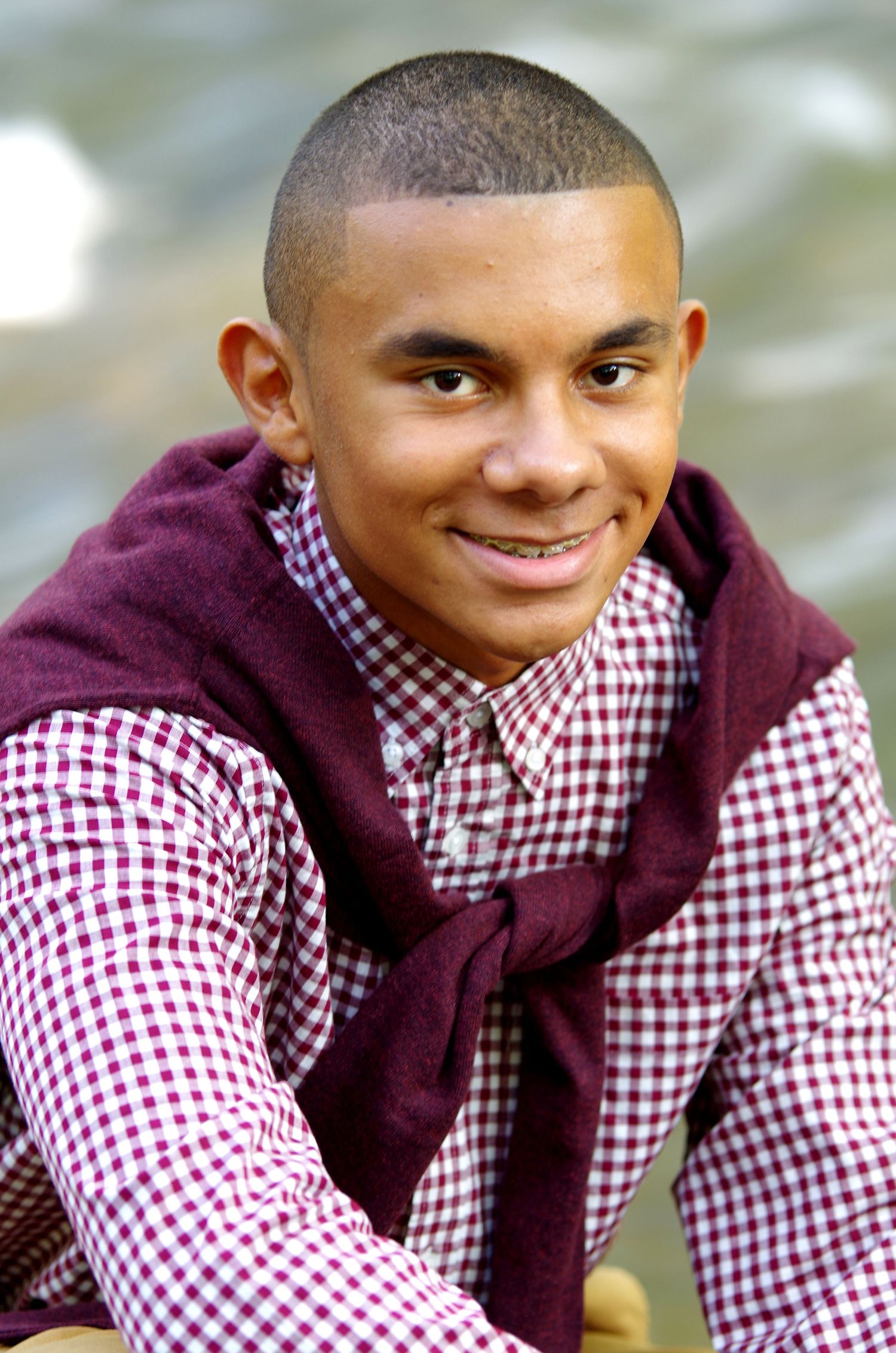 Casual portrait featuring checkered shirt and burgundy sweater with natural backdrop.