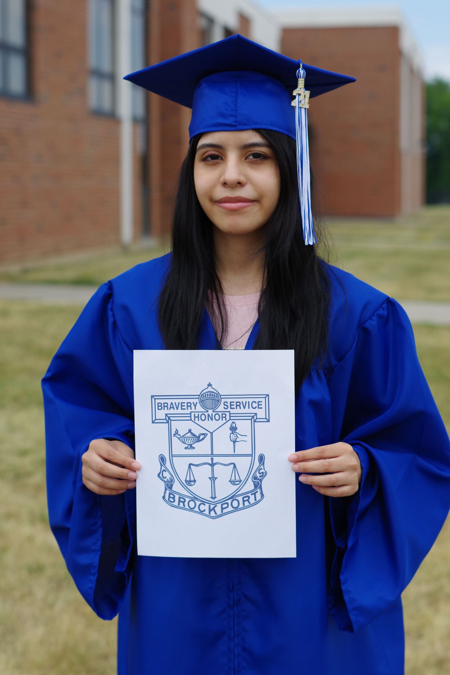 Graduate in royal blue cap and gown holding up a university crest emblem on school grounds.