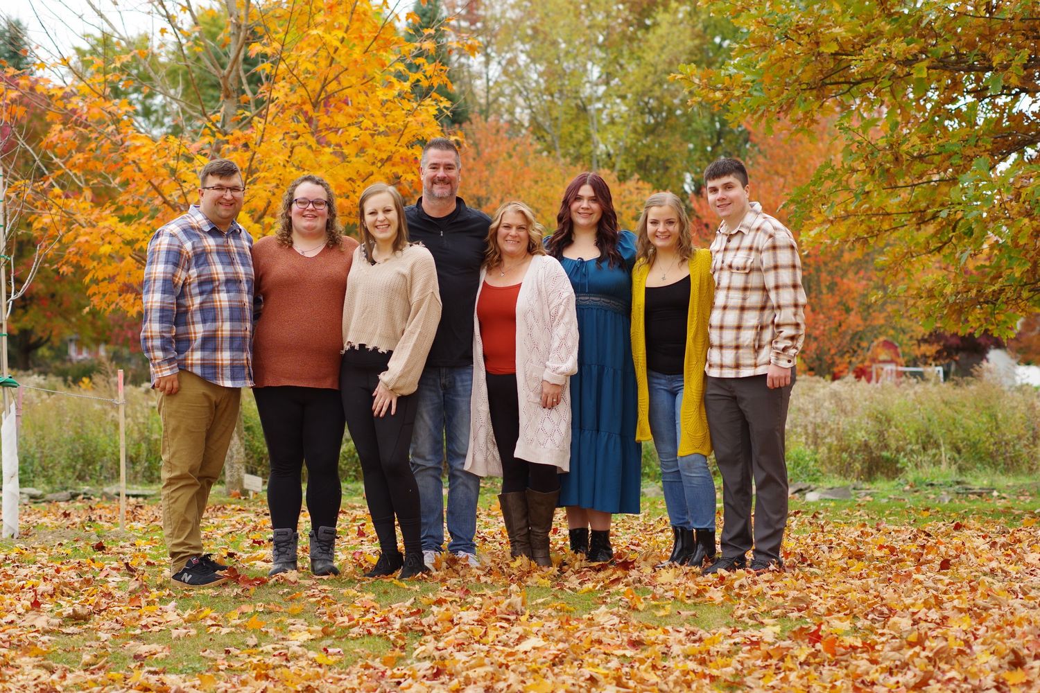 Family of eight posing outdoors among vibrant fall foliage and colorful autumn leaves on the ground.