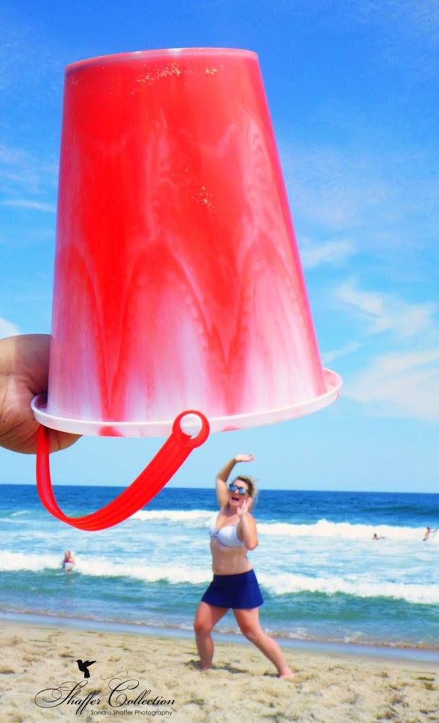Beach photo with red bucket in foreground creating forced perspective with beachgoer in background.