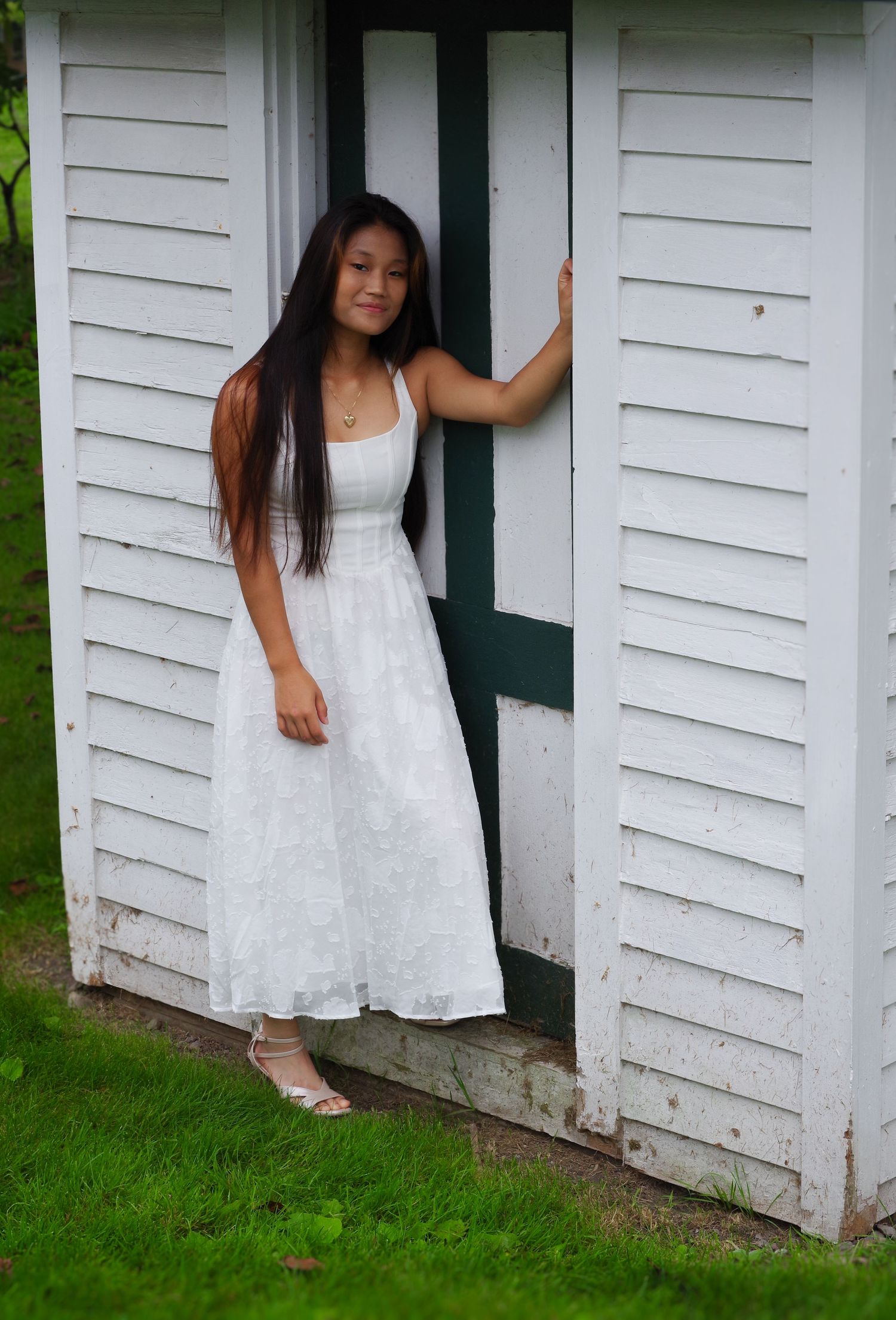 Figure in white dress poses in doorway of rustic white building with green trim.