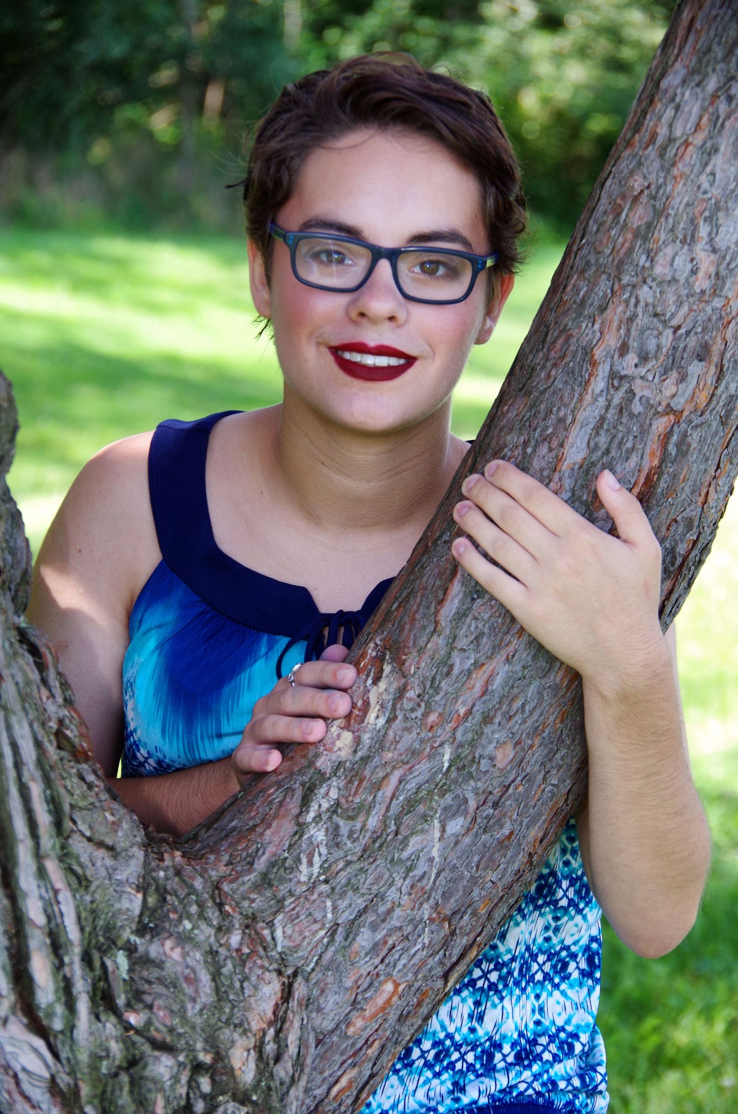 A person wearing glasses and blue dress poses playfully by a tree trunk in a sunny park.