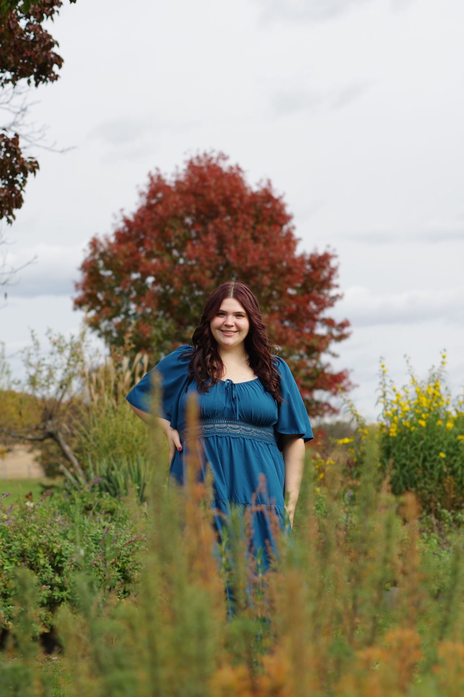 Woman in blue dress standing among tall garden plants with red maple tree behind.