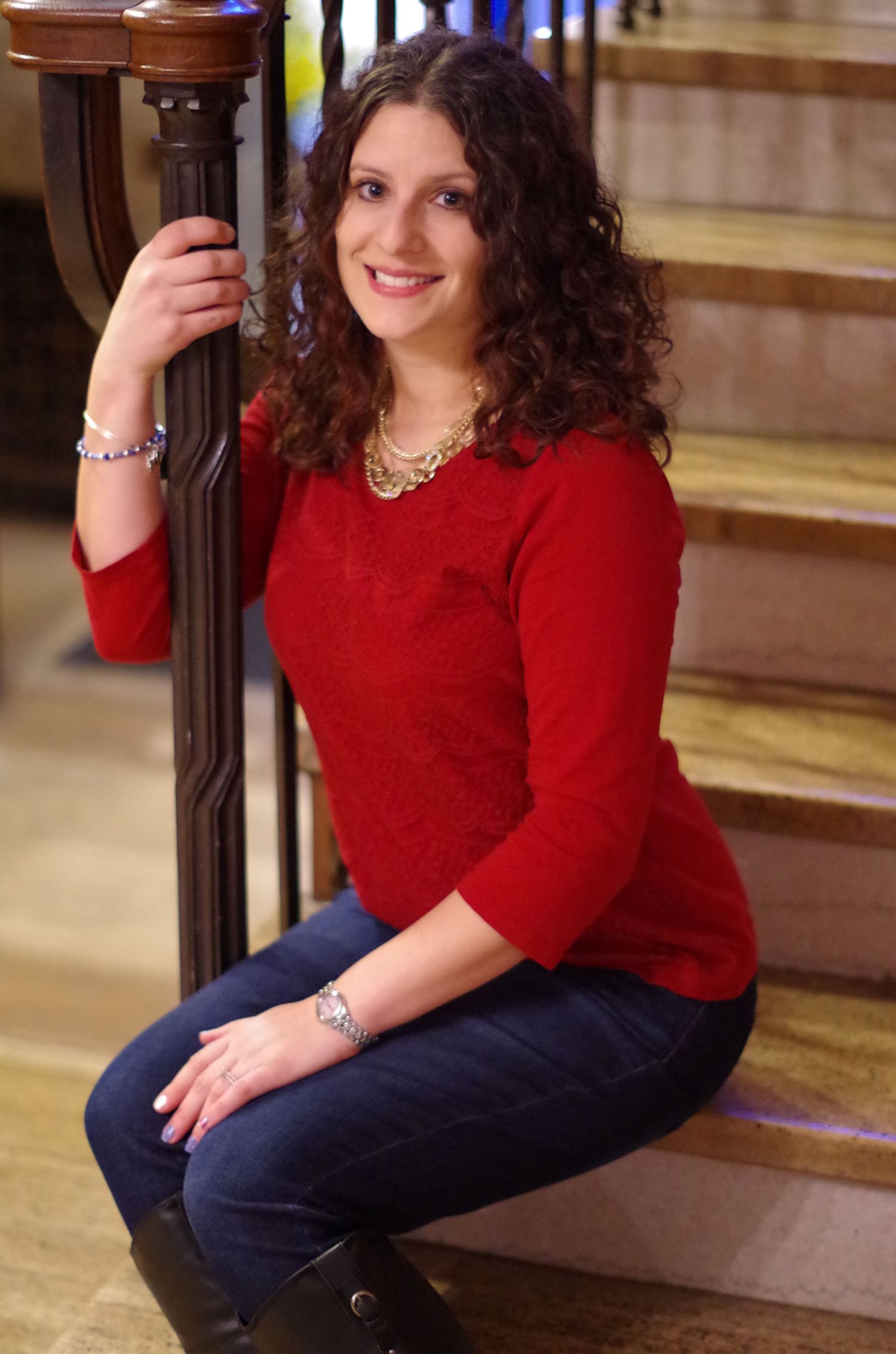 Person in red sweater sitting on wooden stairs with ornate railing while wearing statement jewelry.