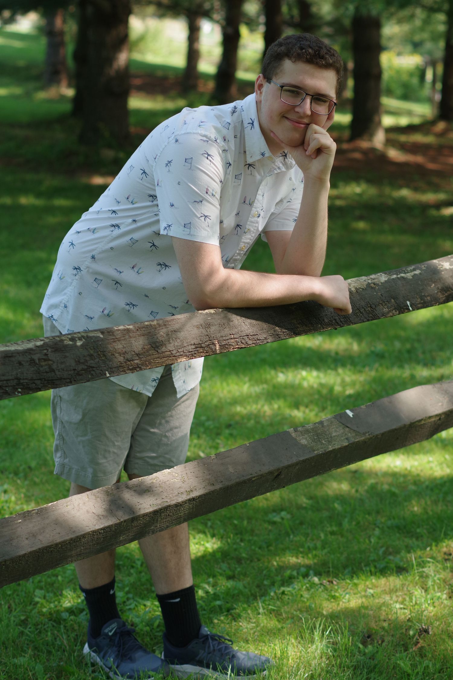 Casual poses leaning on wooden fence wearing patterned shirt in wooded setting.