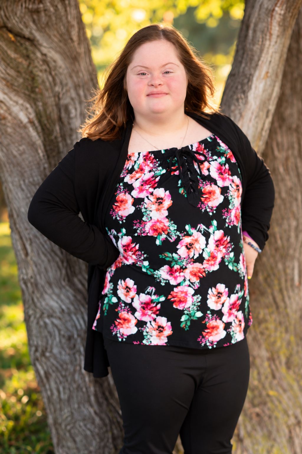 Young woman with Down syndrome smiles outdoors, wearing a floral top and black cardigan, leaning against a tree.