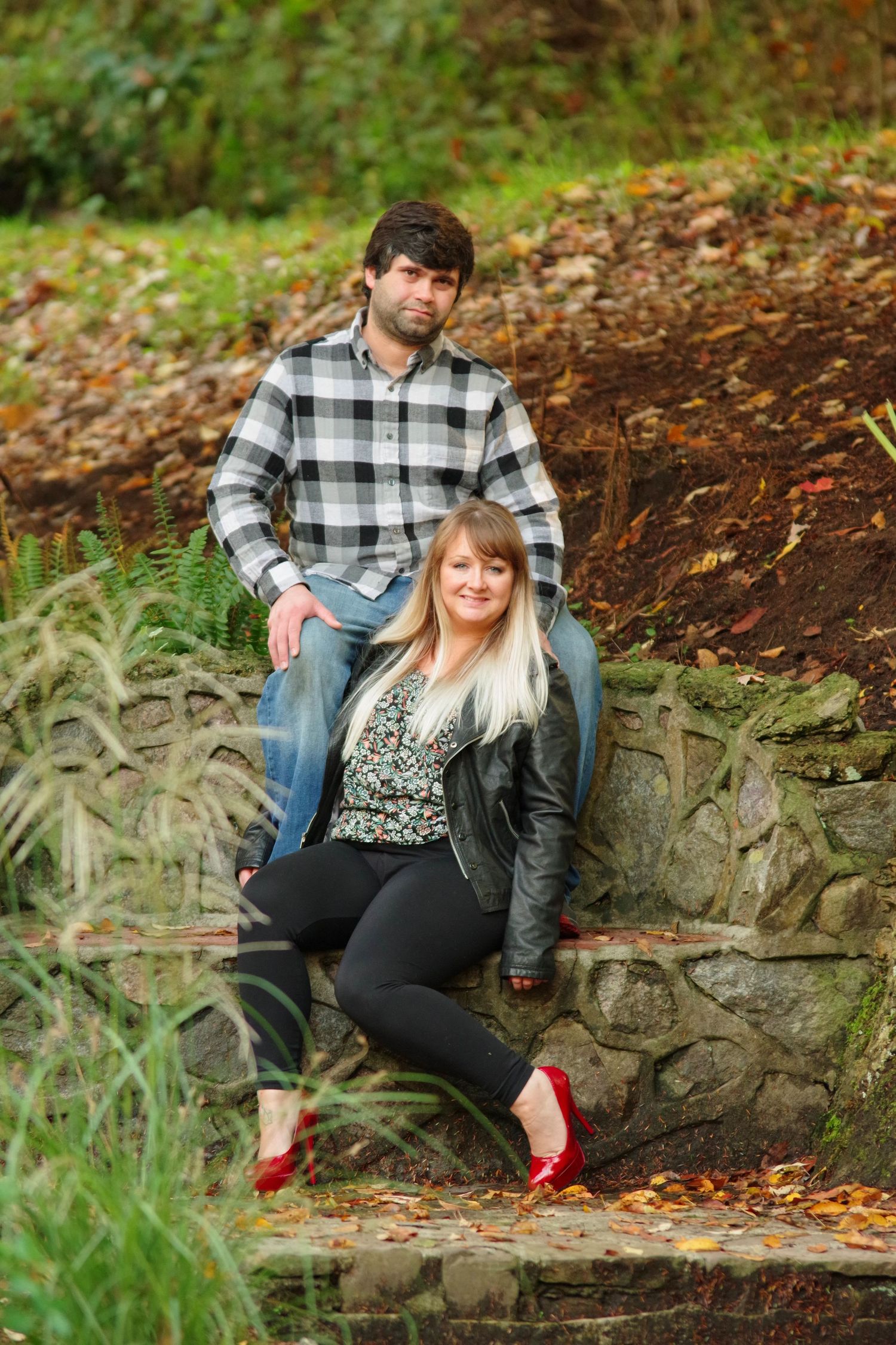 Couple sitting together on rustic stone wall surrounded by autumn foliage in outdoor engagement photos.