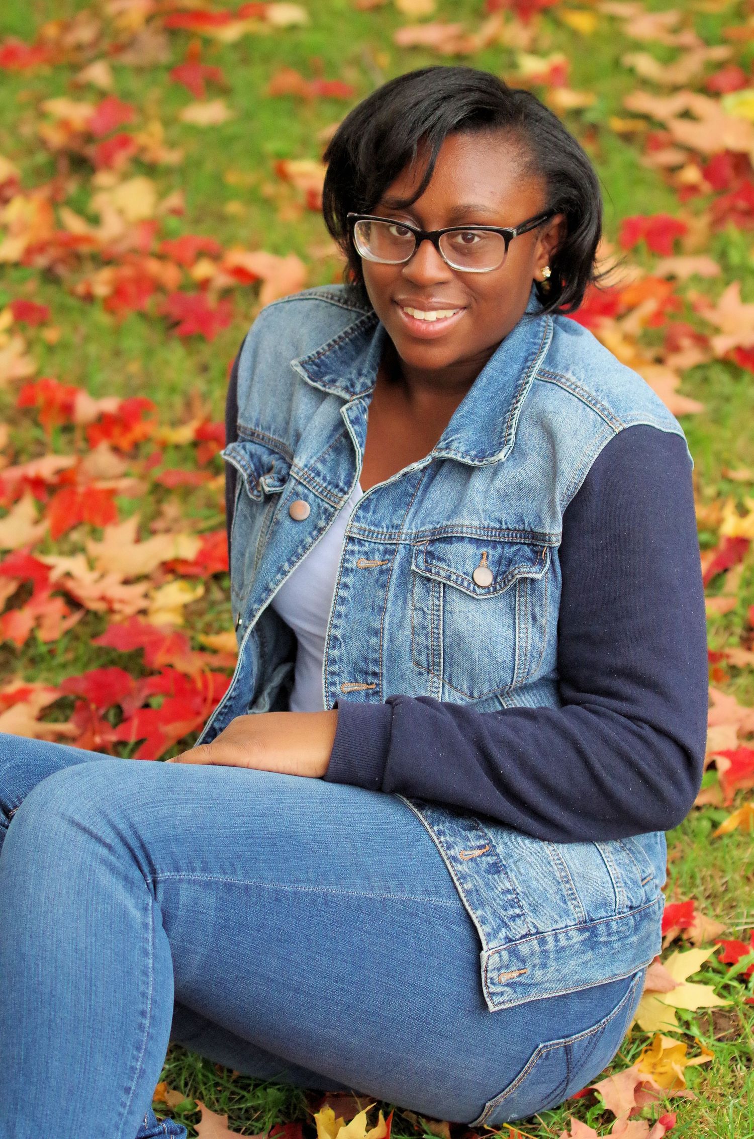 Outdoor portrait seated among fallen red autumn leaves wearing denim vest.