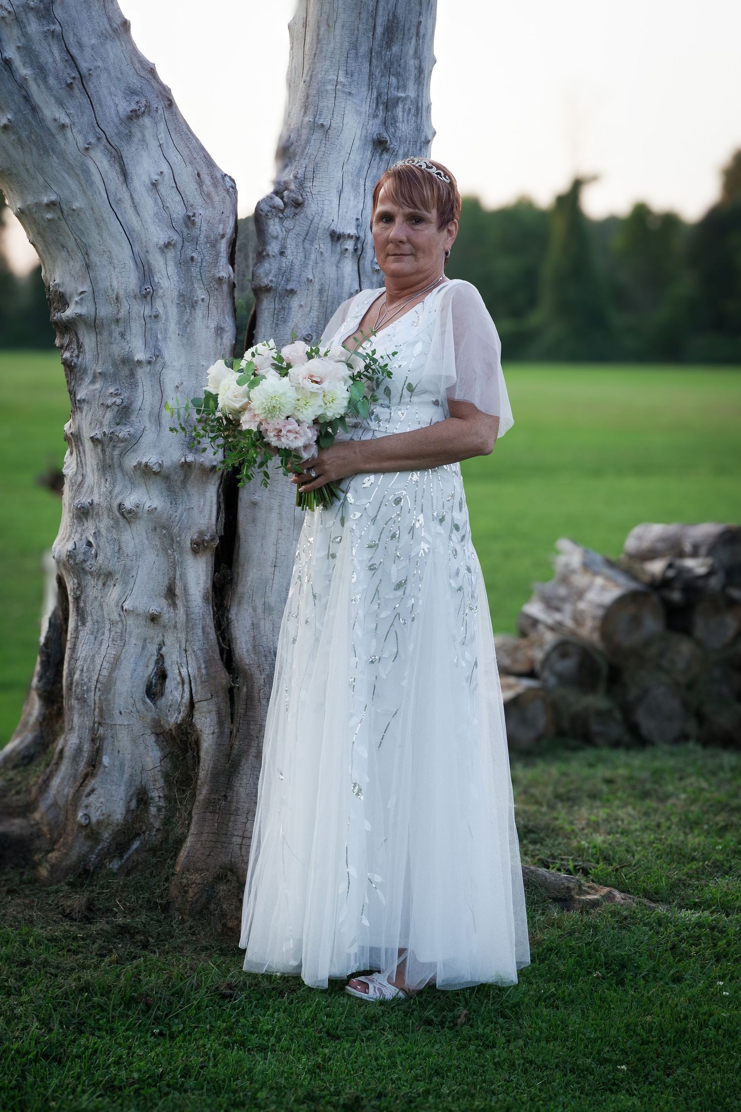 Bride in white lace gown holds bouquet while posing next to white tree trunk.