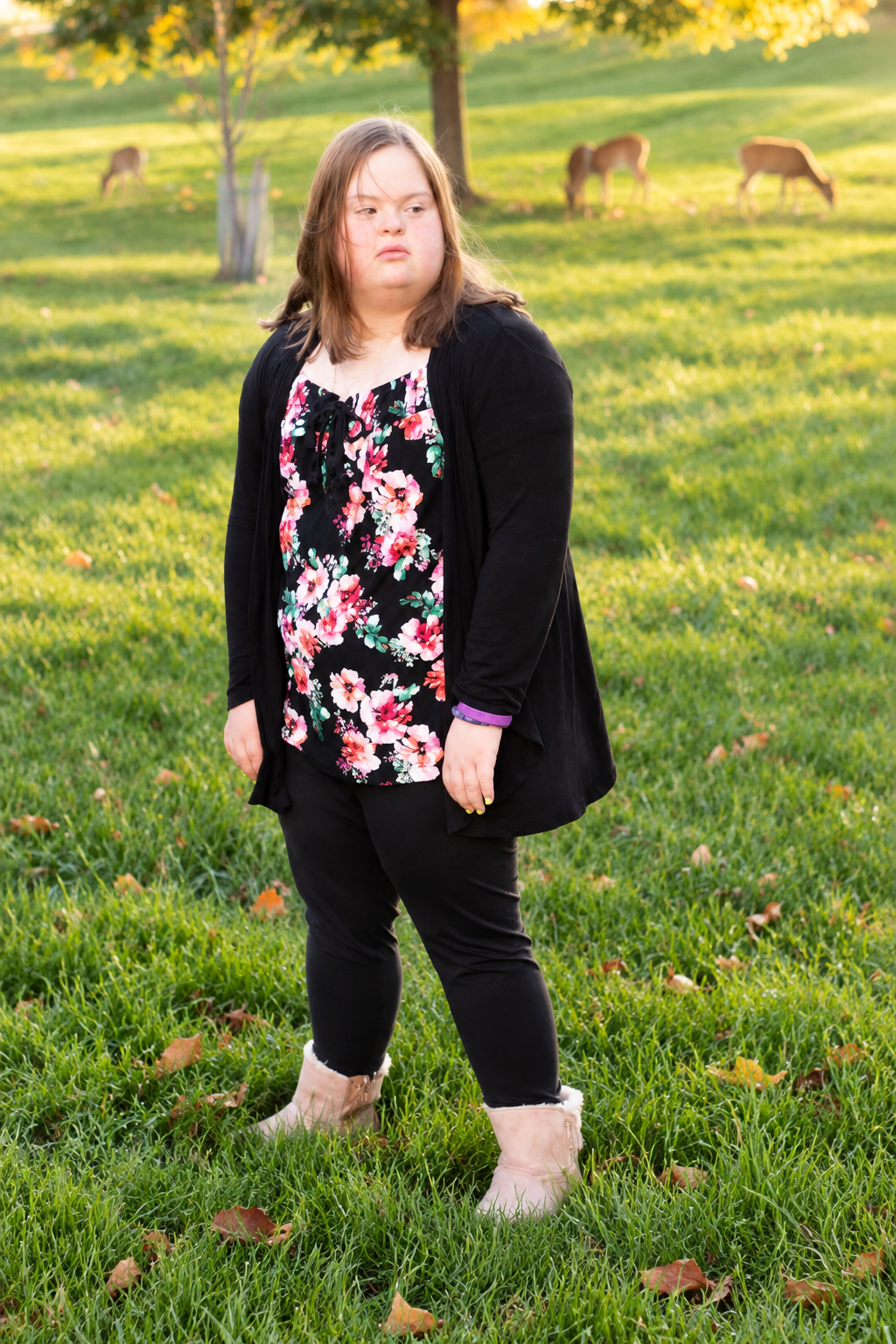 Young woman with Down syndrome poses outdoors in floral top and black cardigan, with deer grazing on green grass behind her.