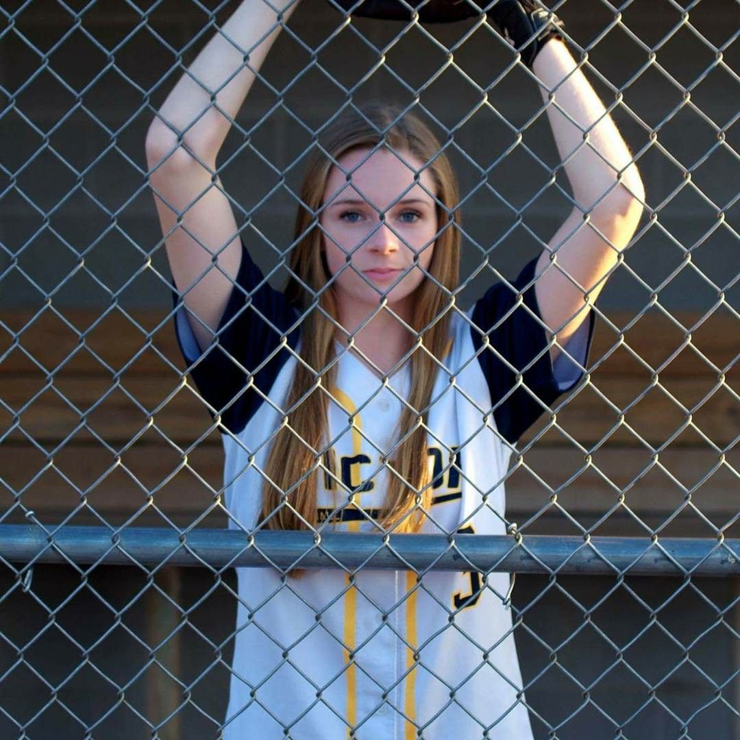 Softball player in uniform poses behind chain link fence during photo session.