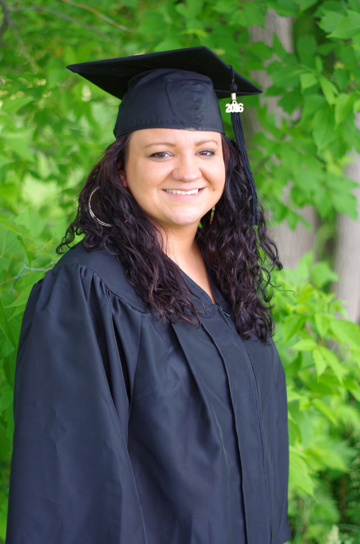 Graduate in traditional black cap and gown poses against lush green foliage.