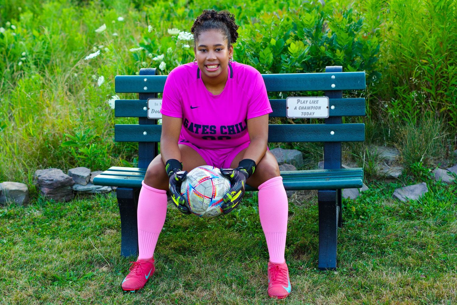A soccer player in bright pink attire sits on a blue bench holding a ball in a sunny outdoor setting.