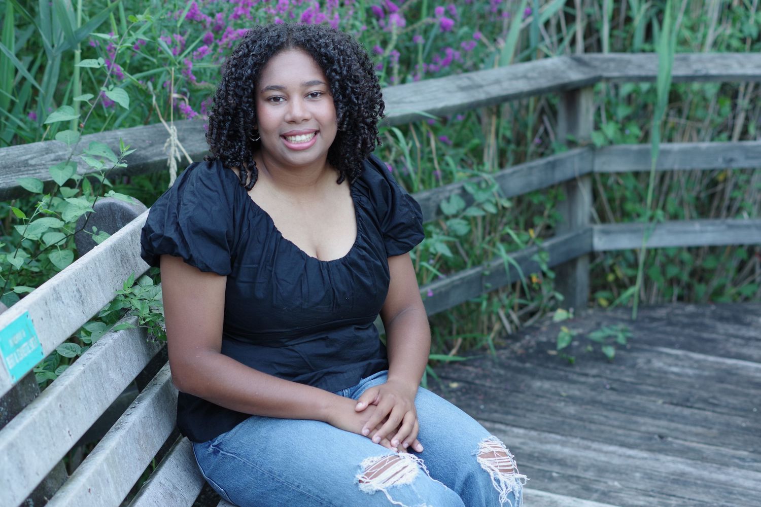 Person sitting on wooden deck surrounded by lush garden greenery wearing a black top and denim jeans.