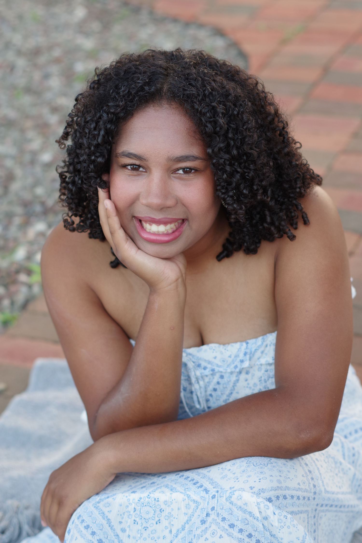 Smiling young woman with curly hair resting chin on hand, wearing a blue strapless dress outdoors.