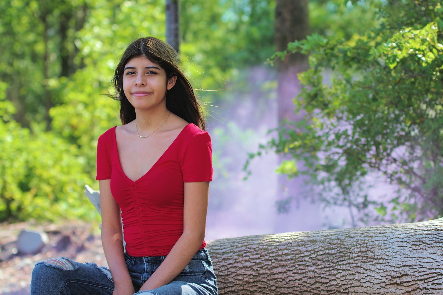Young girl in red top sitting on log in lush green forest with purple smoke in background.