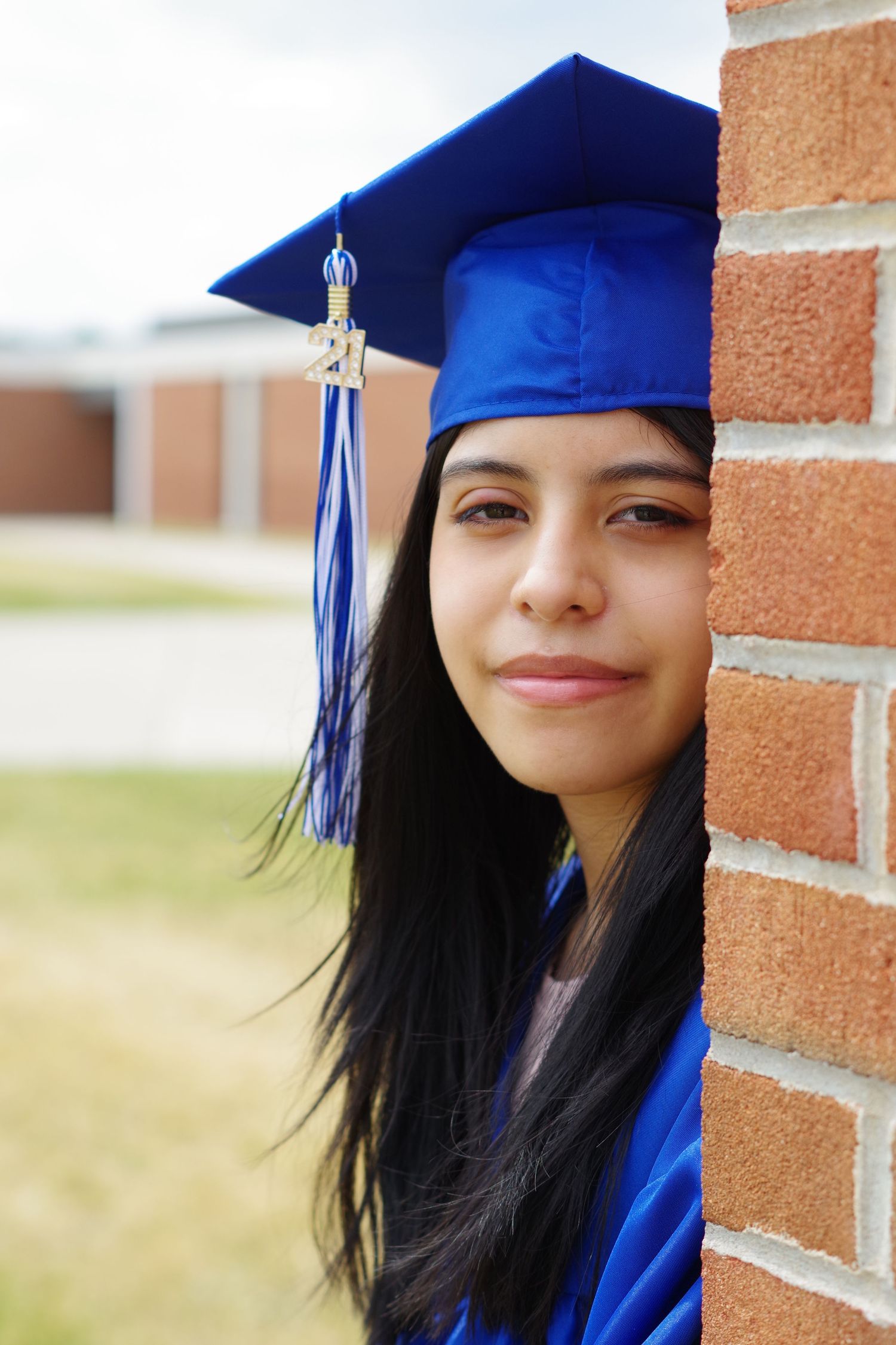 Graduate in bright blue cap and gown poses against brick wall with a gentle smile.