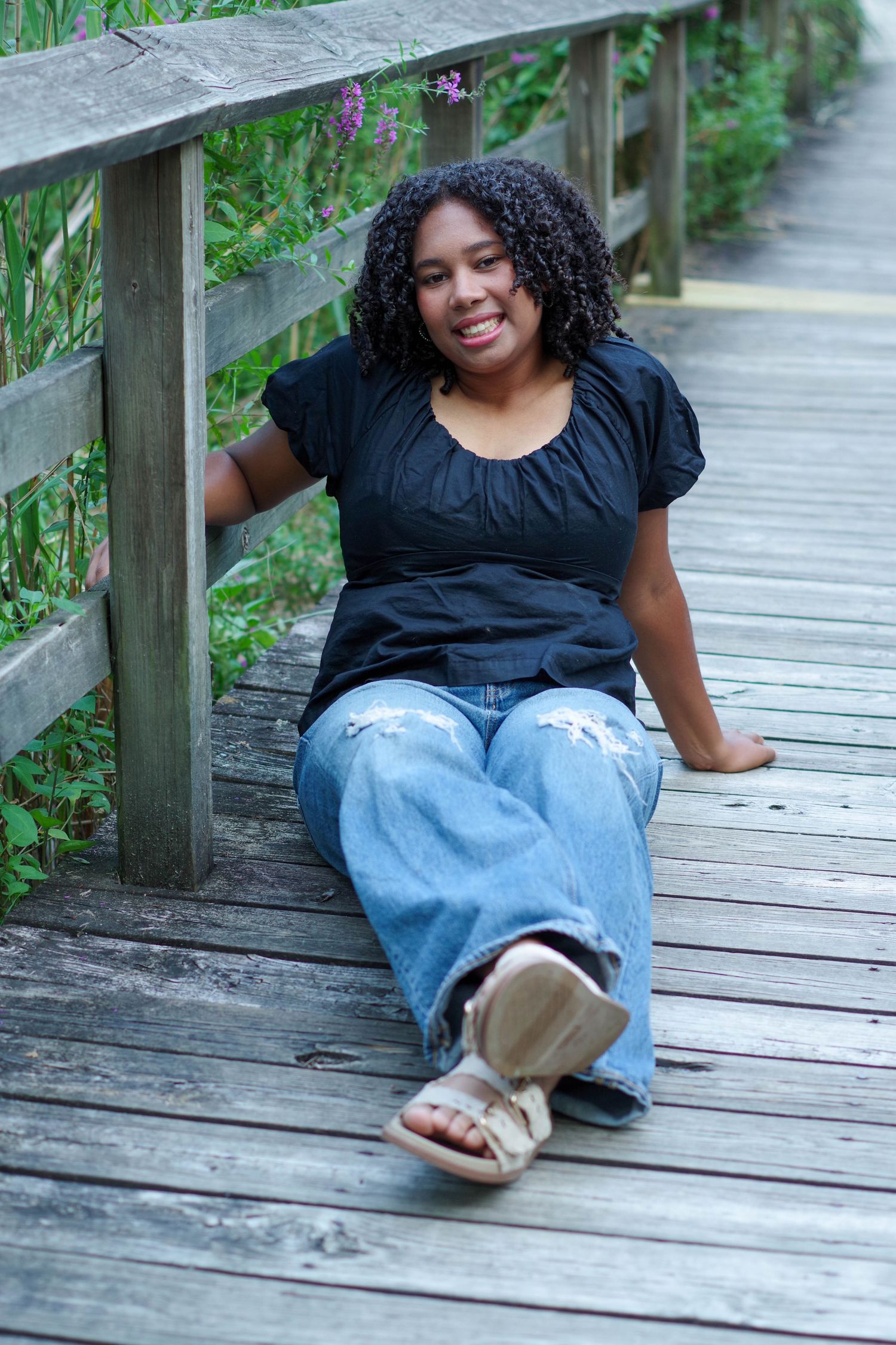 Person in black top and jeans lounges on wooden boardwalk surrounded by greenery.