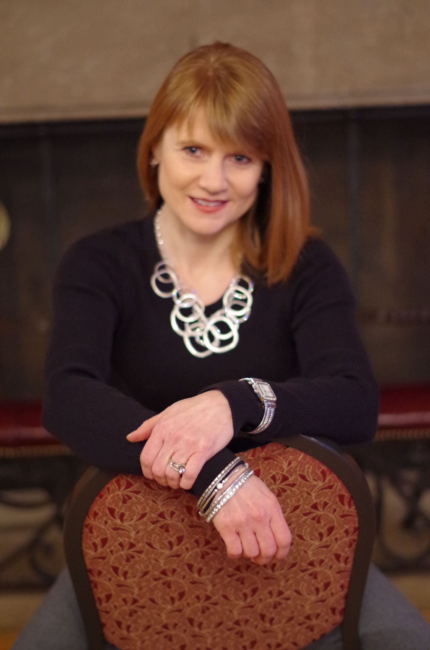 Portrait leaning on upholstered chair wearing black sweater and silver geometric necklace.