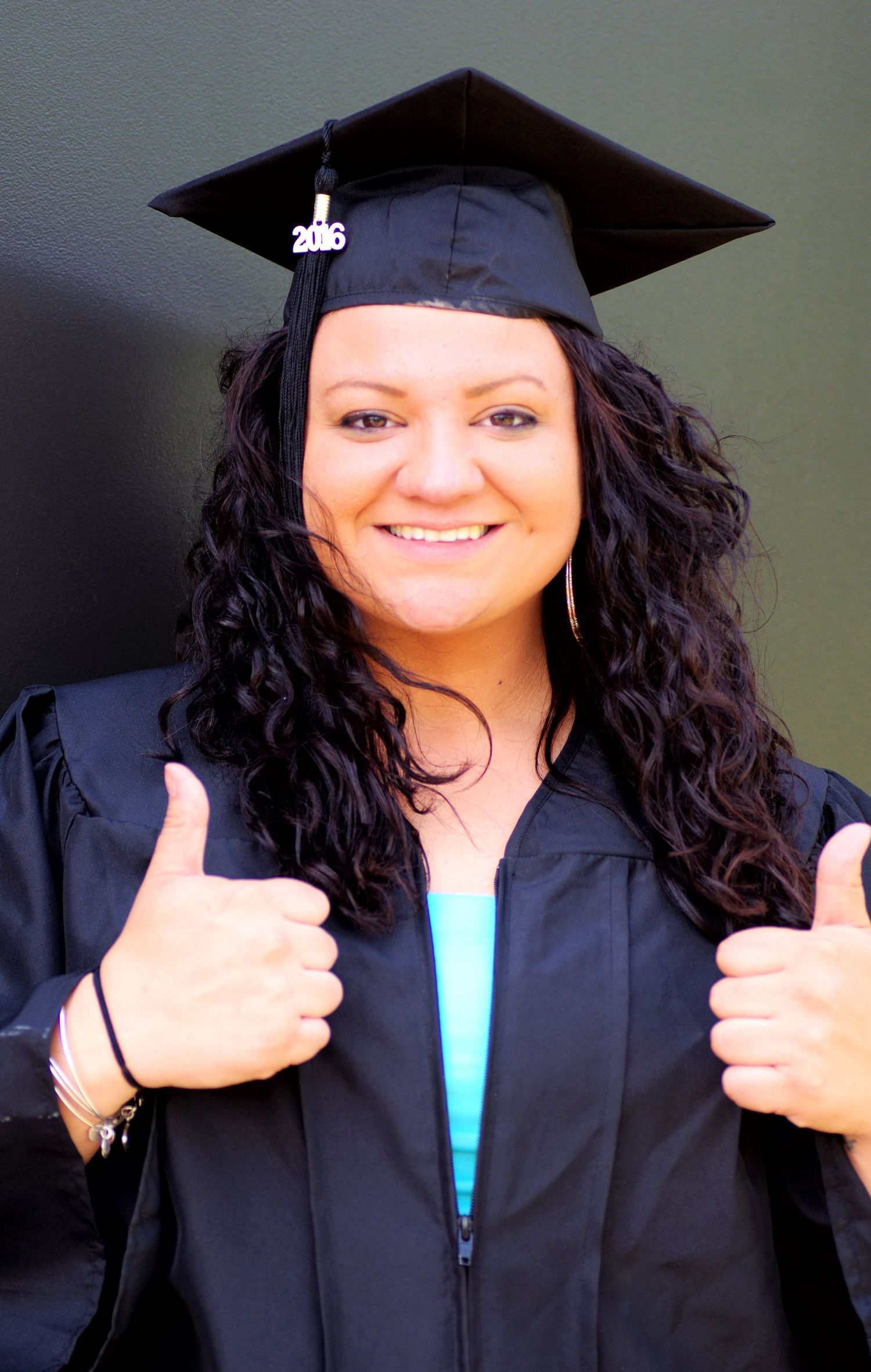 Graduate in black cap and gown gives enthusiastic thumbs up against neutral background.