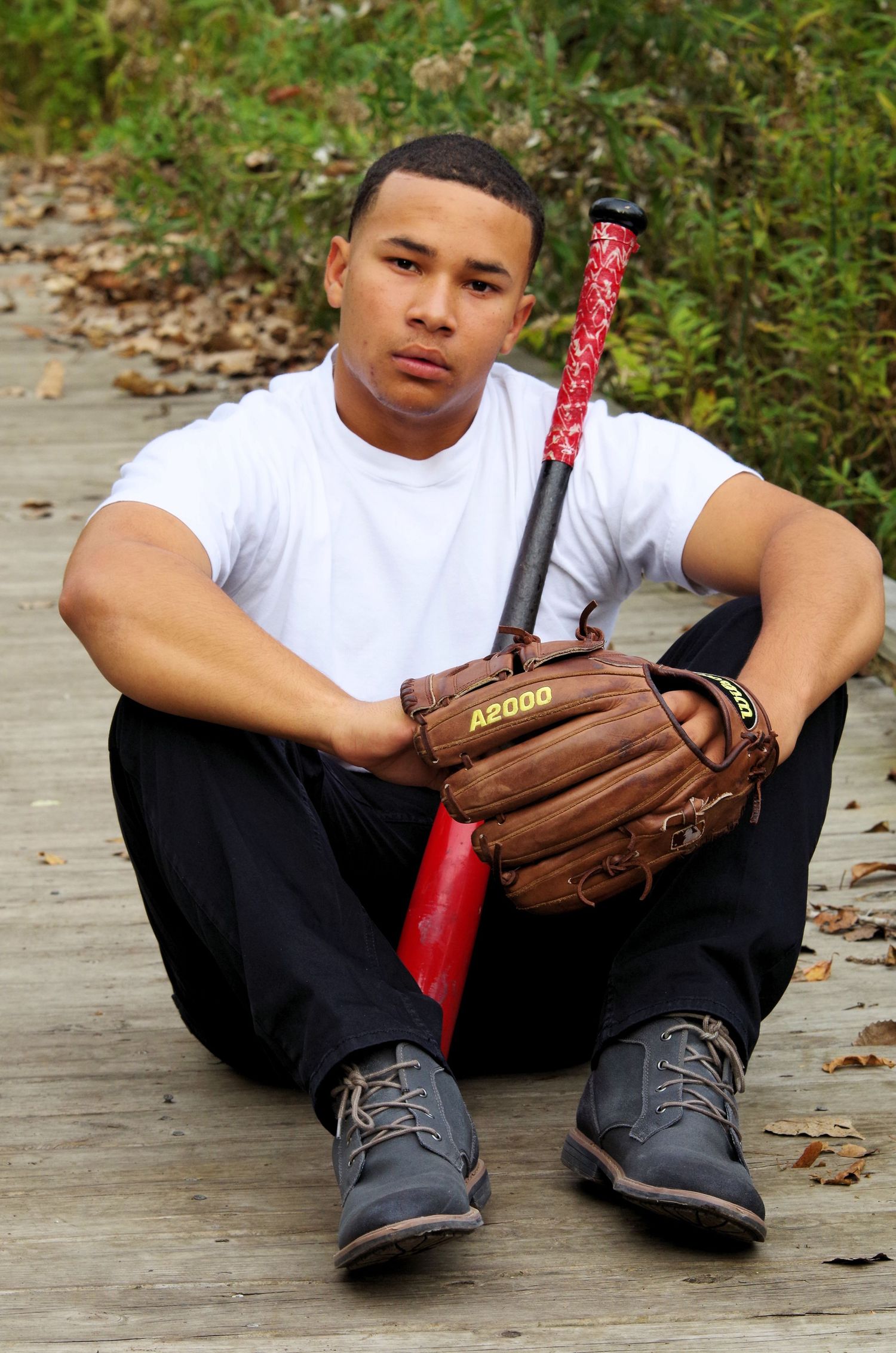Baseball player sitting with bat and glove on concrete path surrounded by autumn leaves.