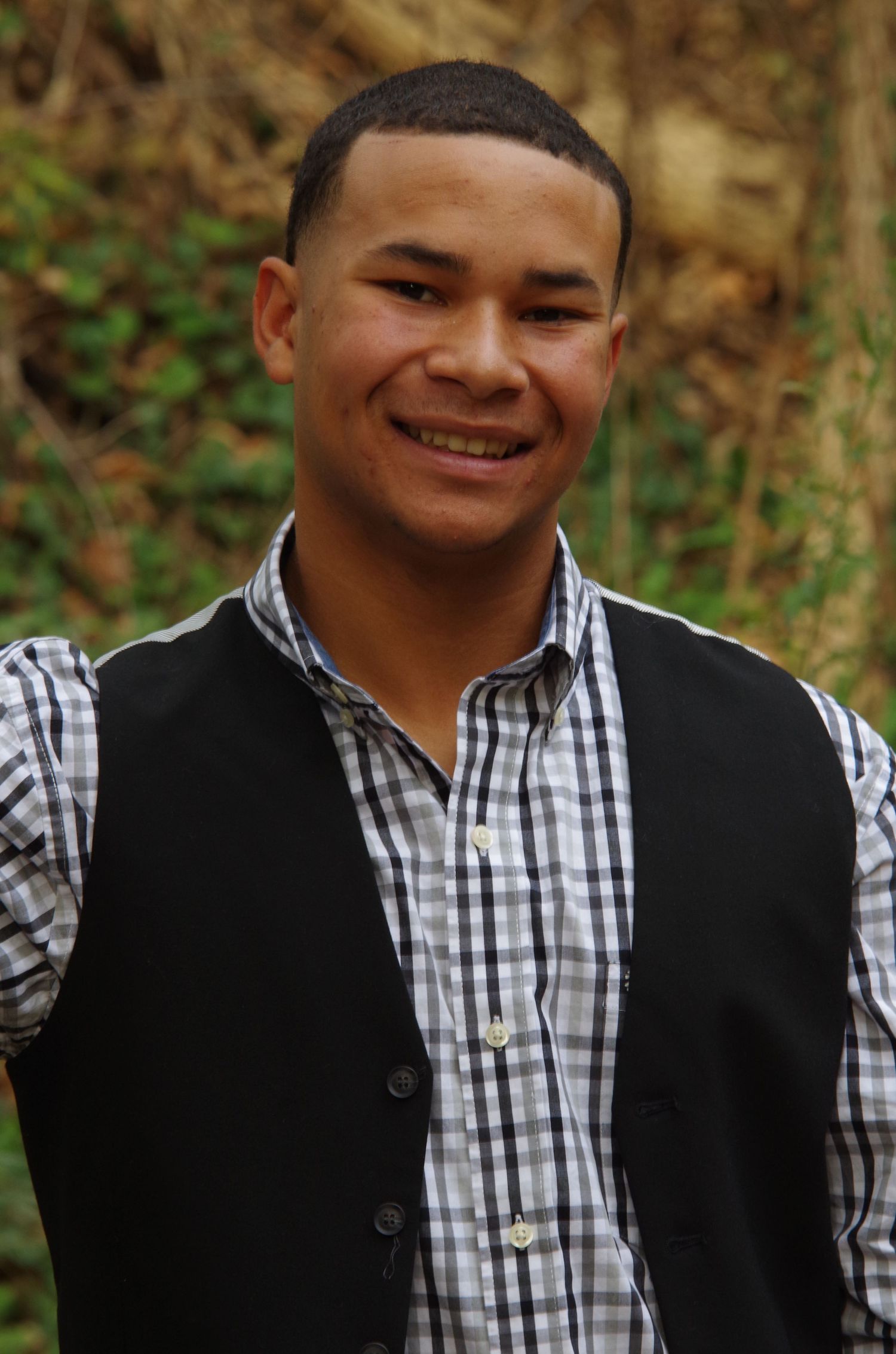 A person in a black vest and plaid shirt poses outdoors with natural foliage in the background.