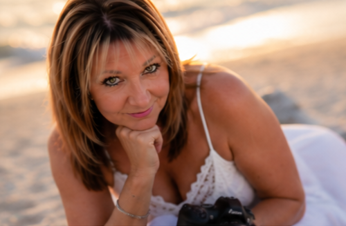 Woman with highlighted hair poses on a sandy beach holding a camera, wearing a white top in warm golden light.