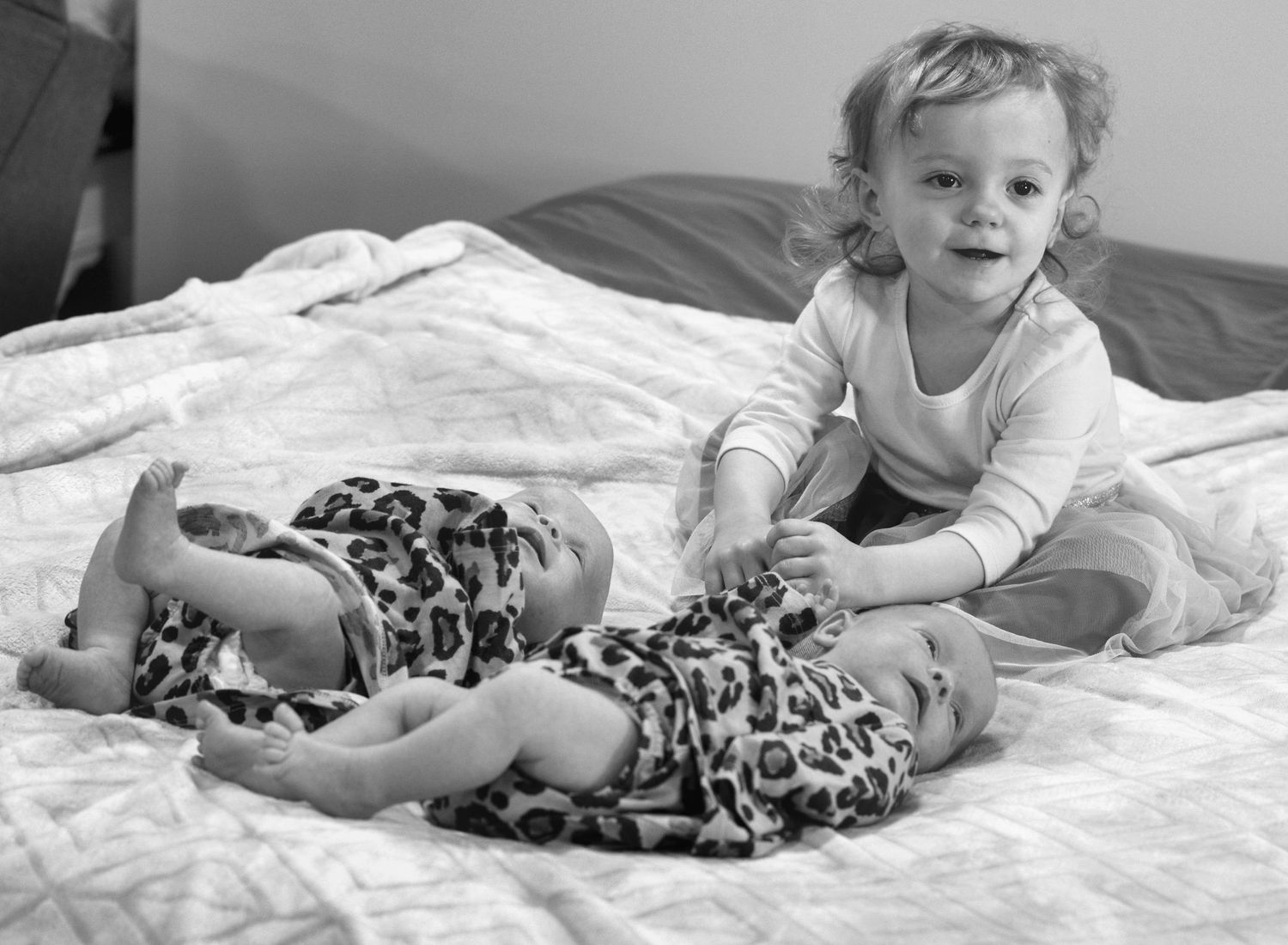 Black and white image of infants in matching outfits on bed with white linens.
