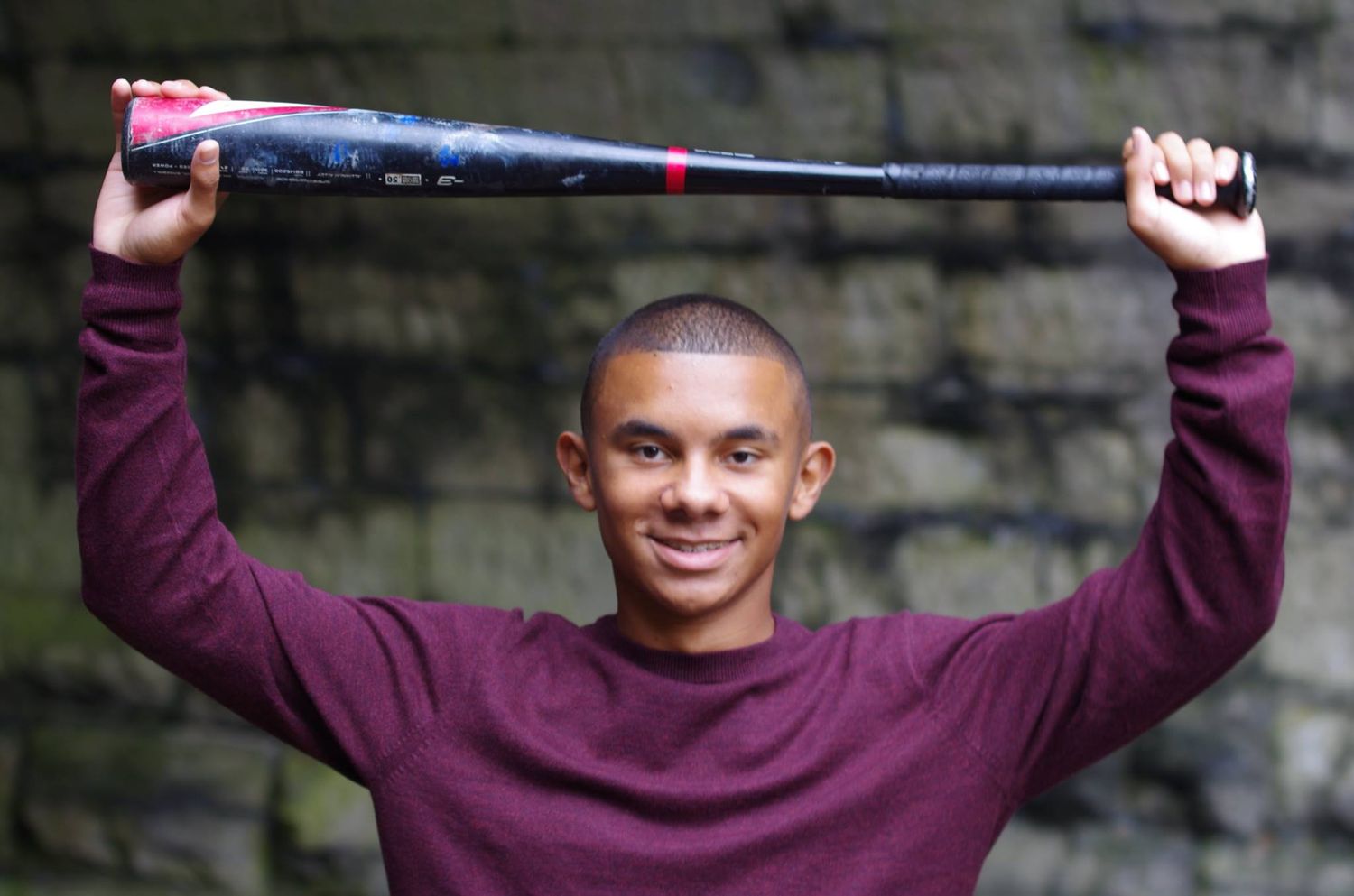 Athletic pose holding baseball bat while wearing burgundy sweater outdoors.